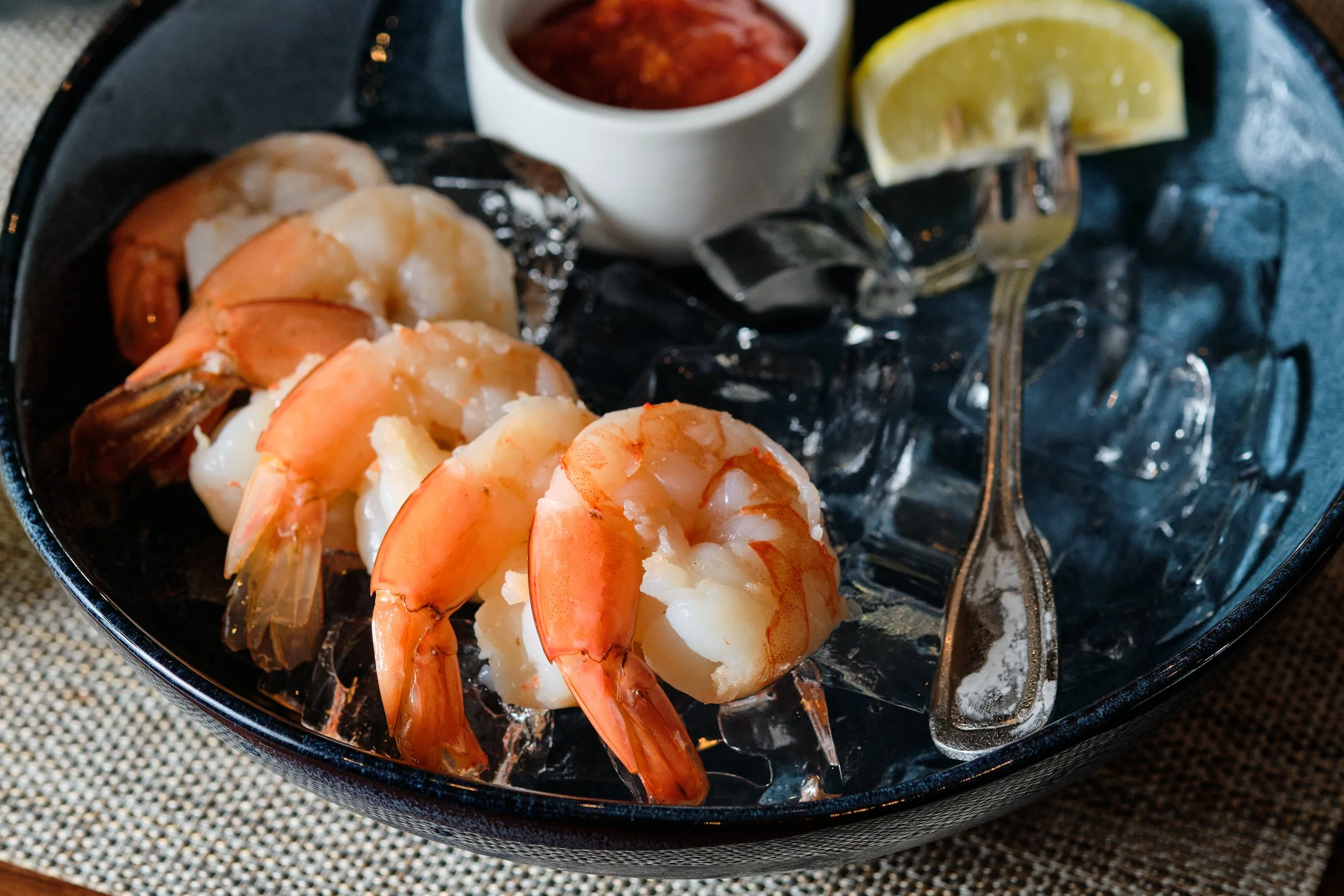Cooked shrimp served on a bed of ice with a lemon wedge, alongside a small container of dipping sauce and a fork.