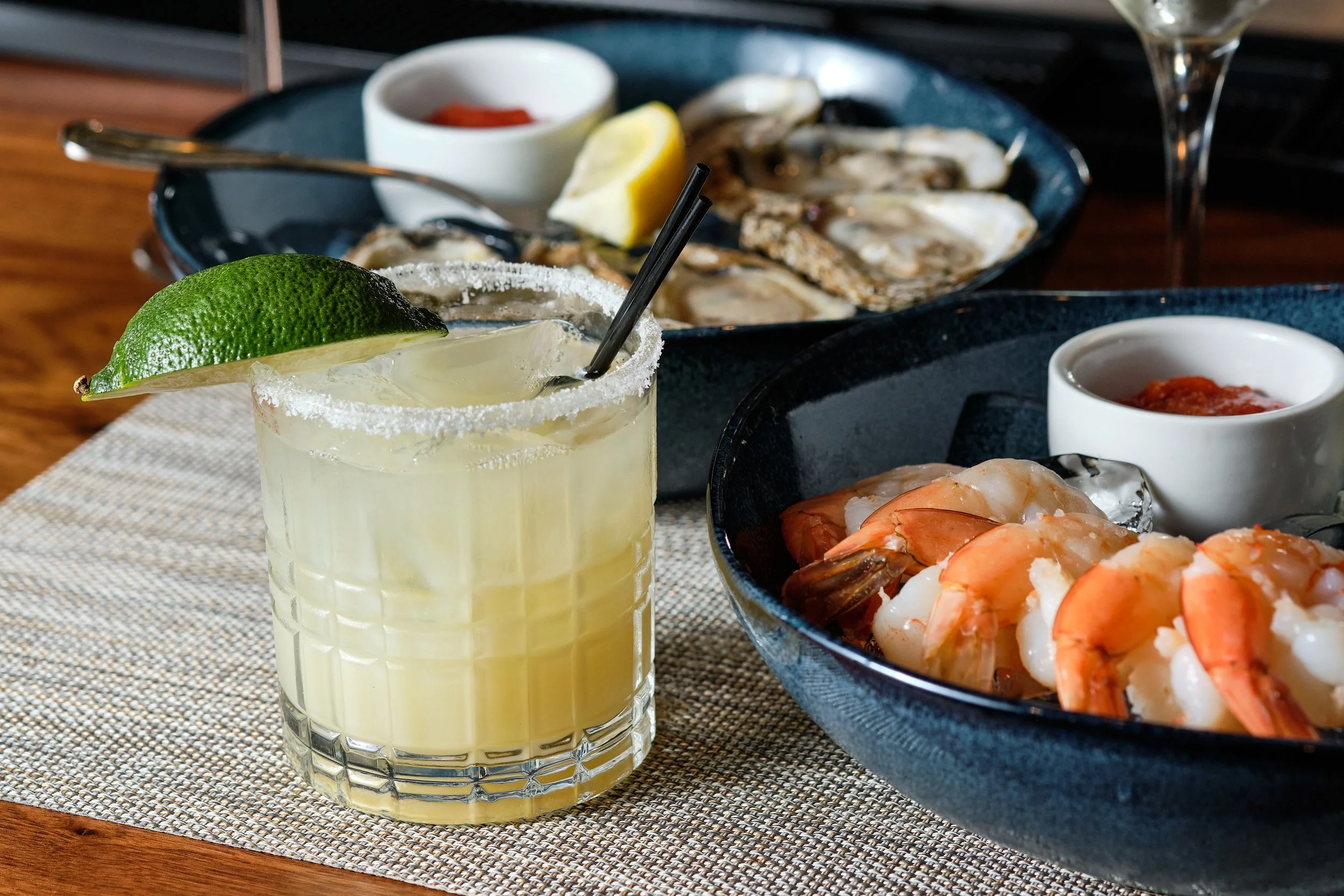 A cocktail with a salted rim, lime wedge, and straw in the foreground. Behind are bowls of seafood, including shrimp and oysters, with sauces and lemon wedges.