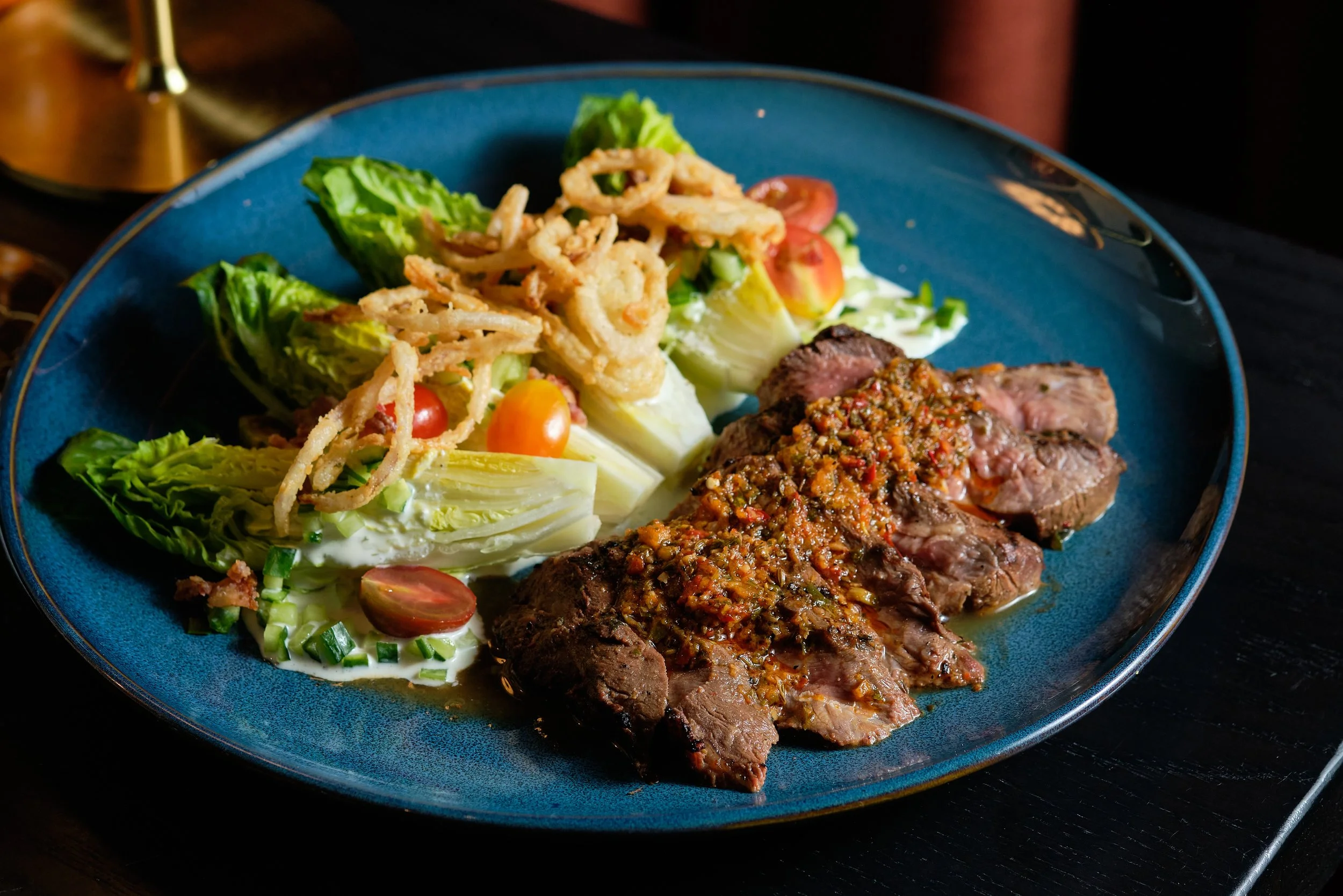 Plate of sliced steak topped with a spicy sauce, side salad with cherry tomatoes, lettuce, and onions, and crispy onion rings.