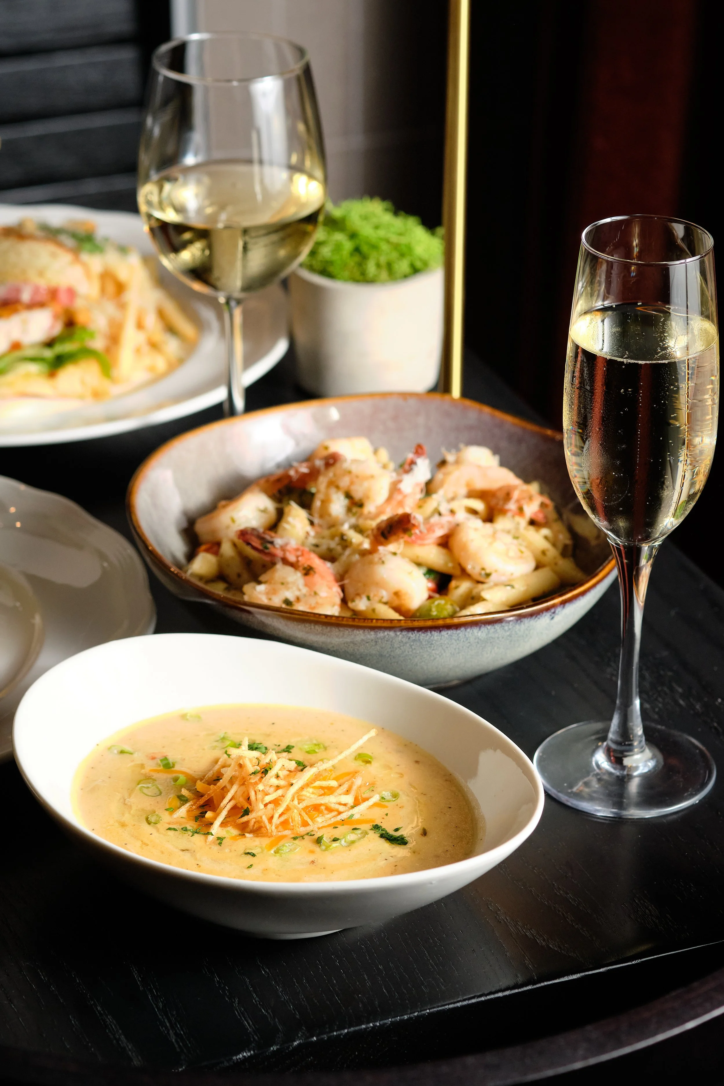 A dining table with a bowl of creamy soup garnished with shredded cheese and chopped green onions, a bowl of shrimp and pasta, and two glasses of white wine.