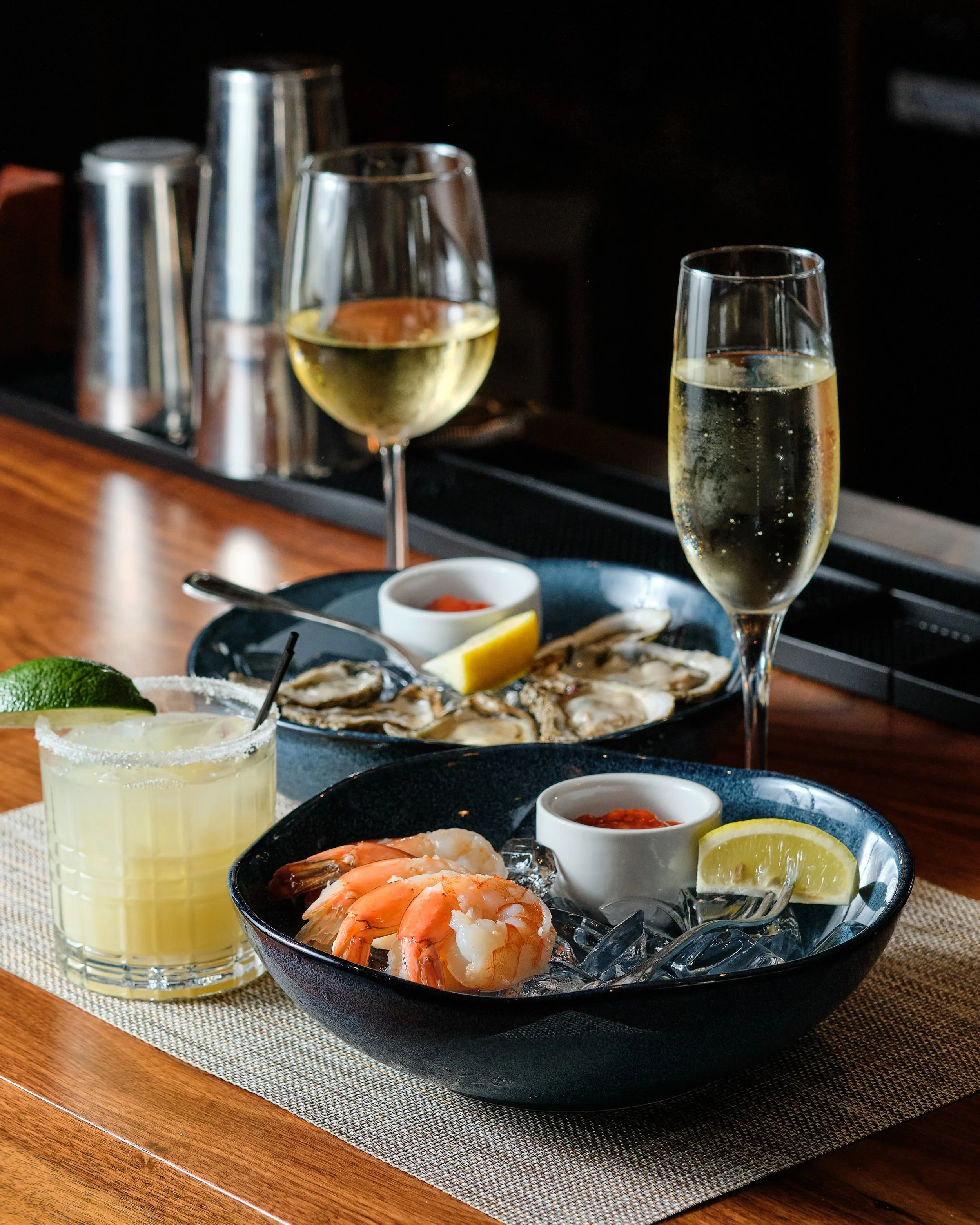 Seafood platter with shrimp, oysters, lemon wedges, and dipping sauces on a table, accompanied by glasses of white wine and a cocktail with lime