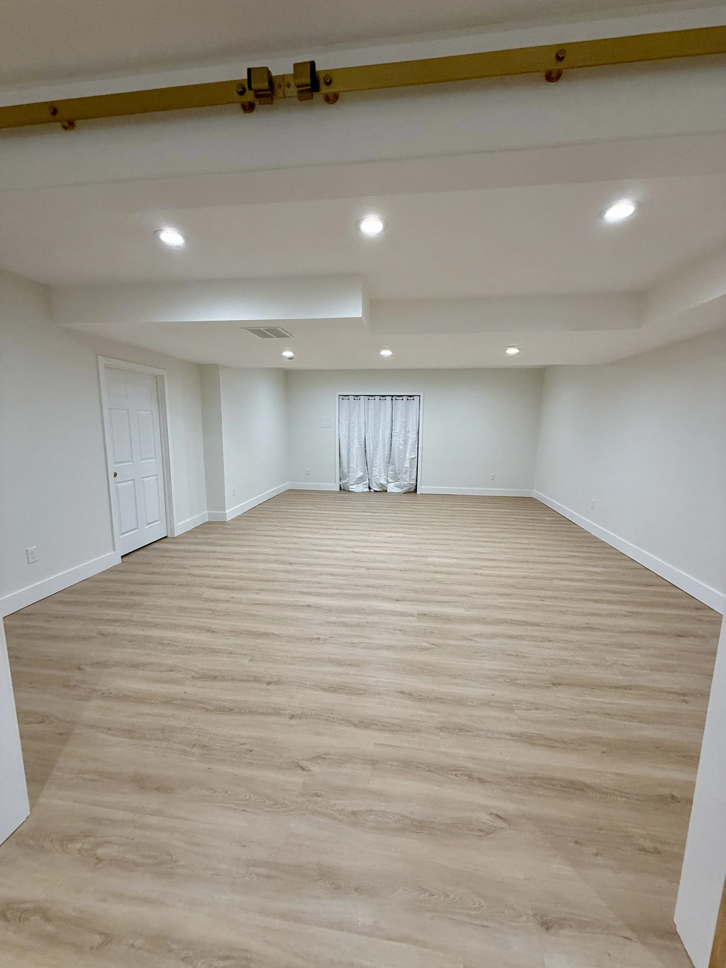 Empty finished basement with light wood flooring, white walls, ceiling lights, a door, and a small window covered with curtains.
