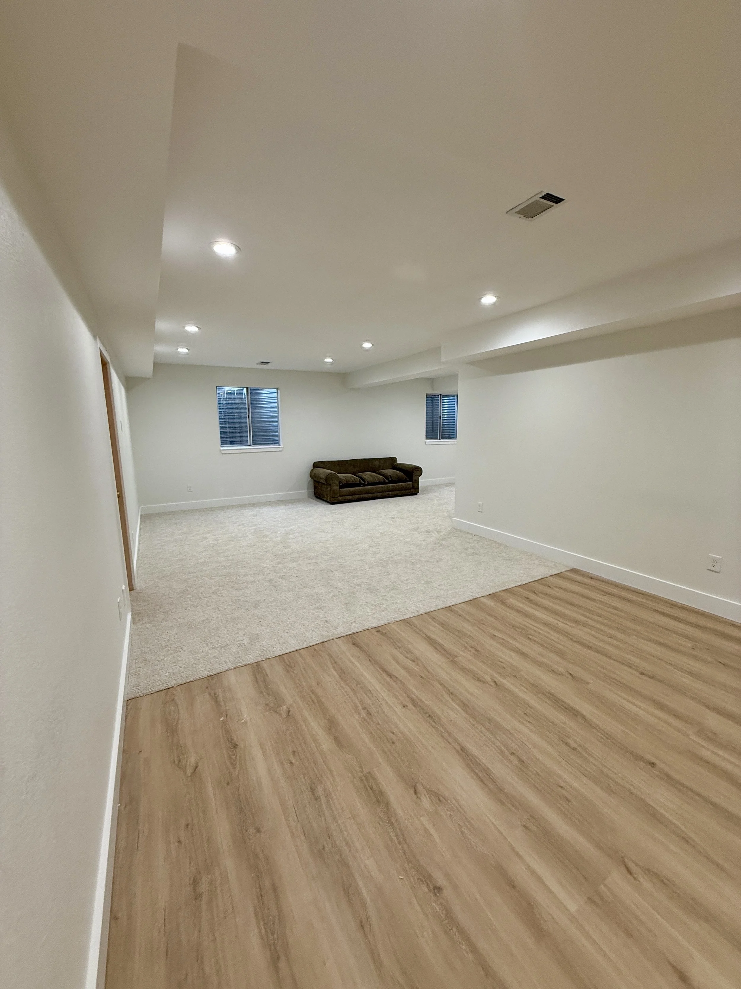 Empty basement room with white walls, wooden and beige carpeted flooring, three small windows, ceiling lights, and a brown couch.