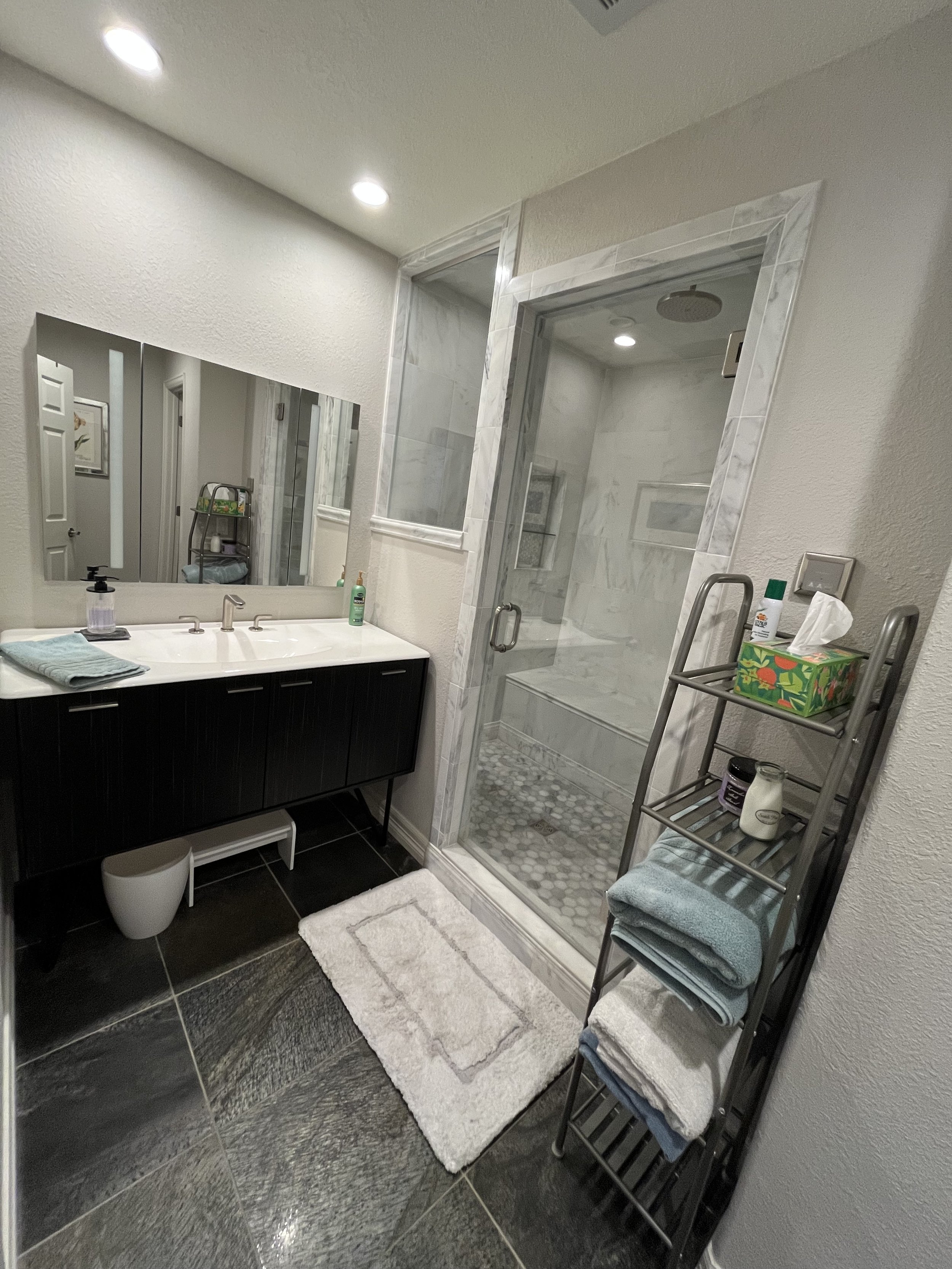 A modern bathroom featuring a black vanity with a sink and a mirror, a walk-in shower with a glass door and marble tiles, and a metal shelf with towels and toiletries.