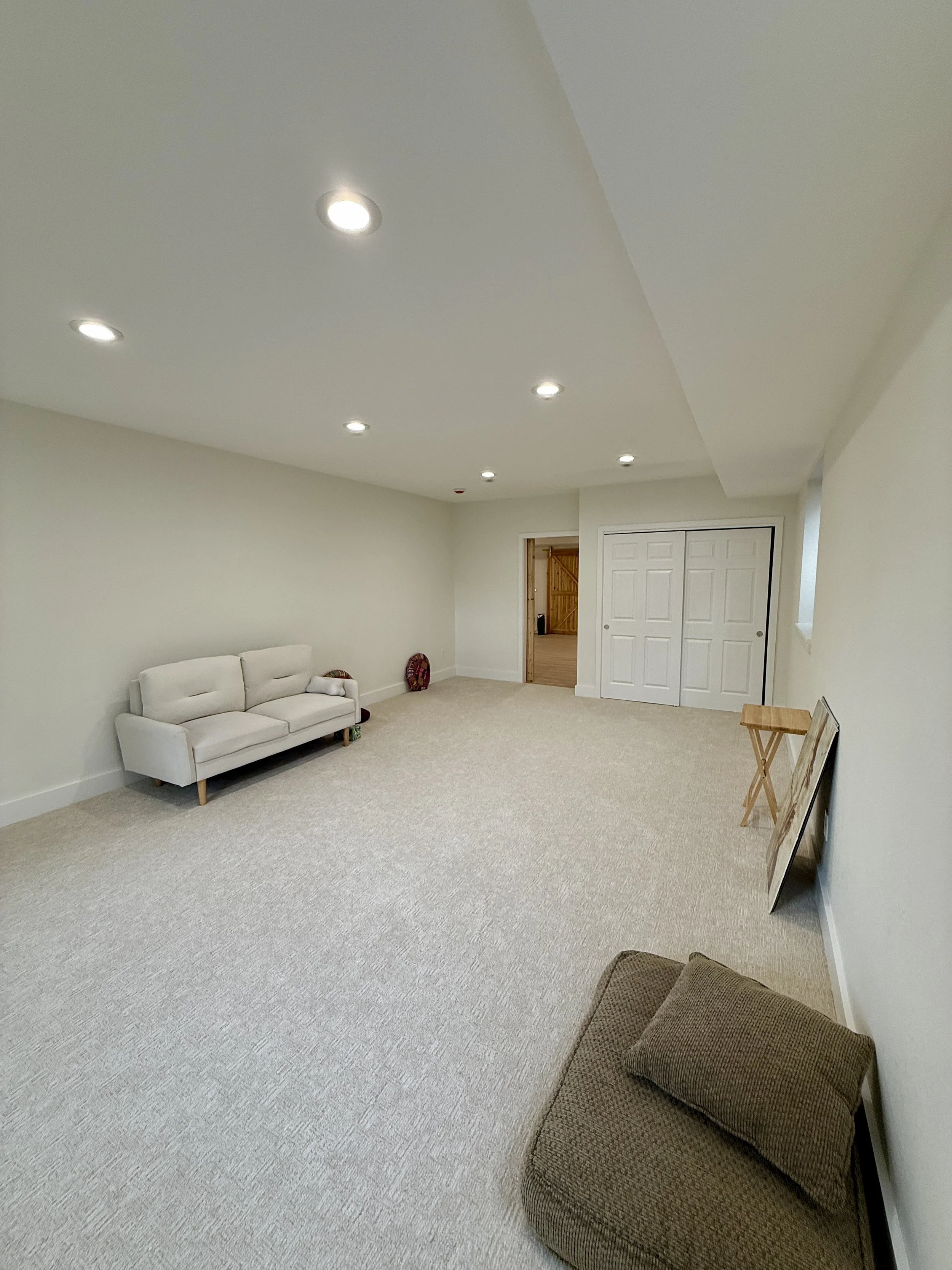 Empty living room with beige carpet, white walls, ceiling lights, a white couch on the left, a brown ottoman in the foreground, a folding table against the right wall, and a closed closet door on the back wall.