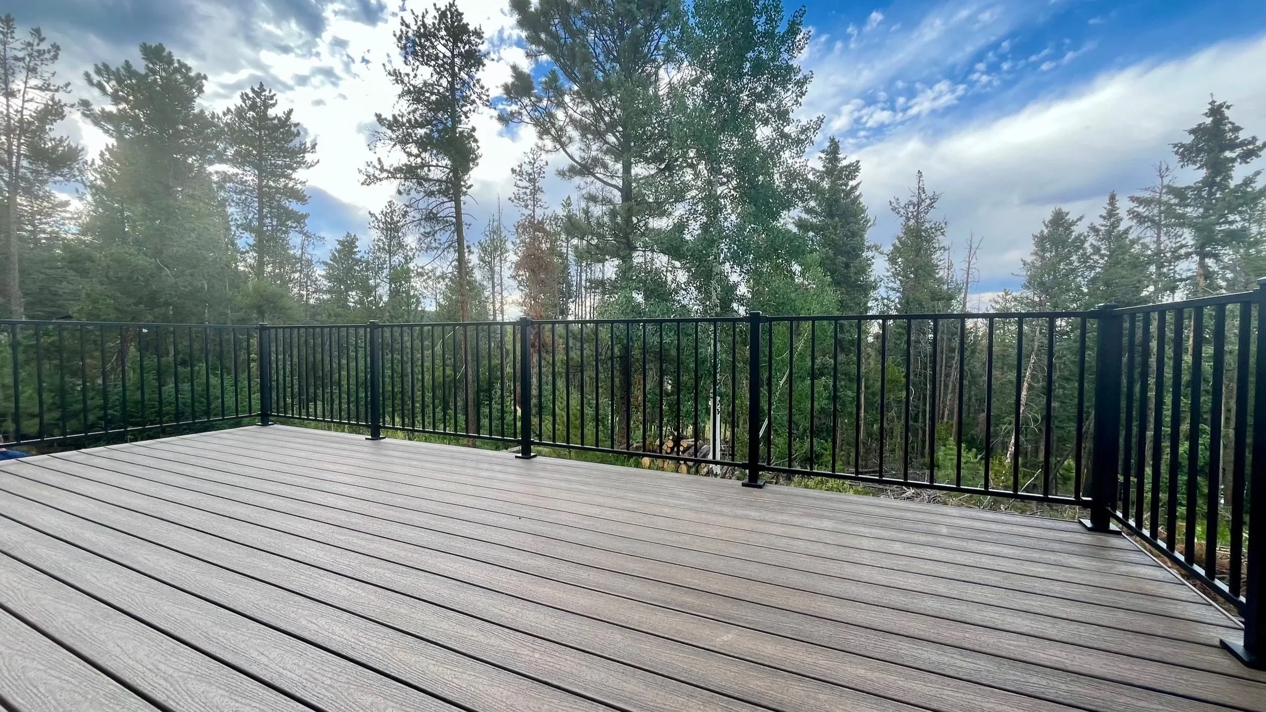 View of a wooden deck with black metal railing overlooking a forested area with tall pine trees under a partly cloudy sky.