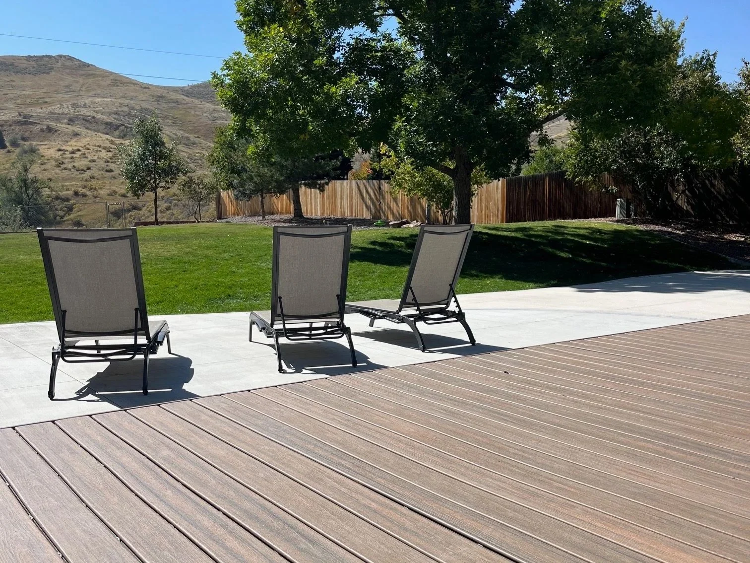 Three lounge chairs on a concrete patio with grass and trees, fenced yard, hillside in background, sunny day.