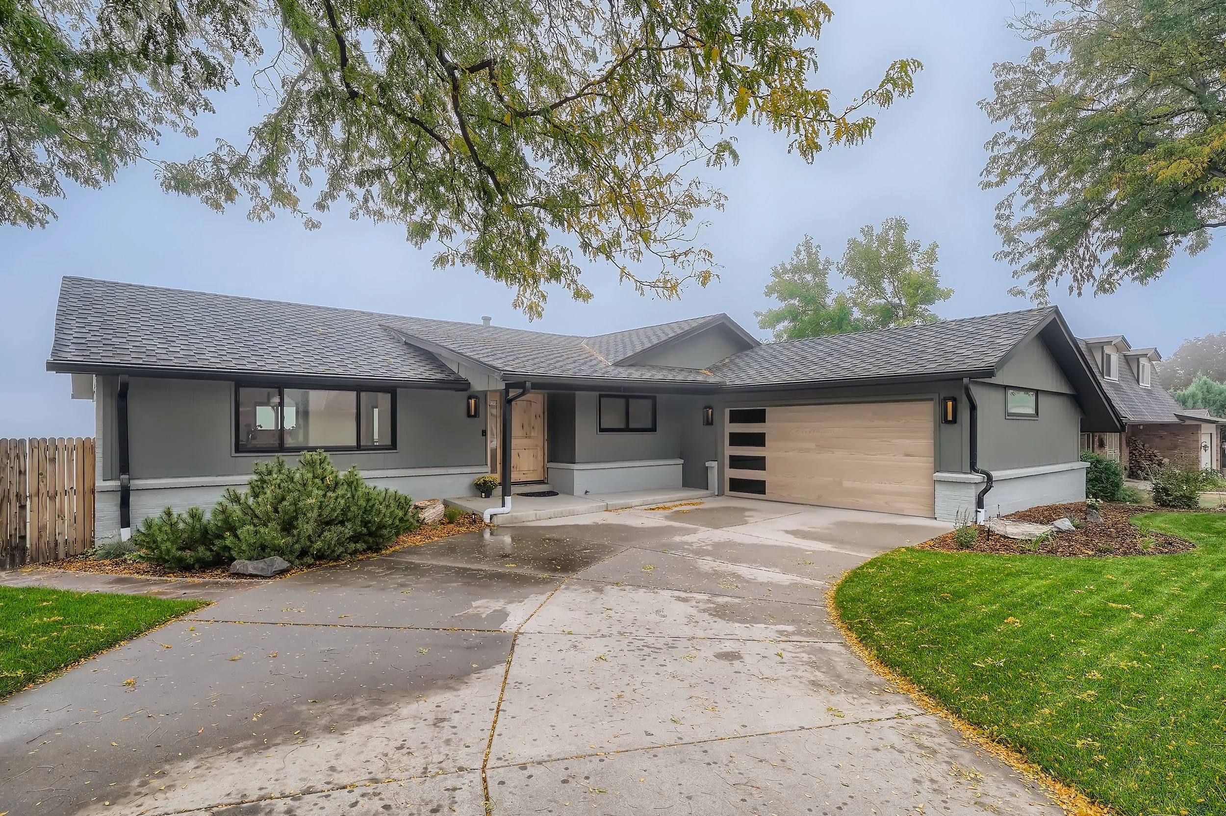 Front view of a modern suburban house with gray siding, black trim, a wooden front door, a two-car garage with a wooden panel door, and a curved concrete driveway. The house is surrounded by green lawn, trees, and landscaping with rocks.