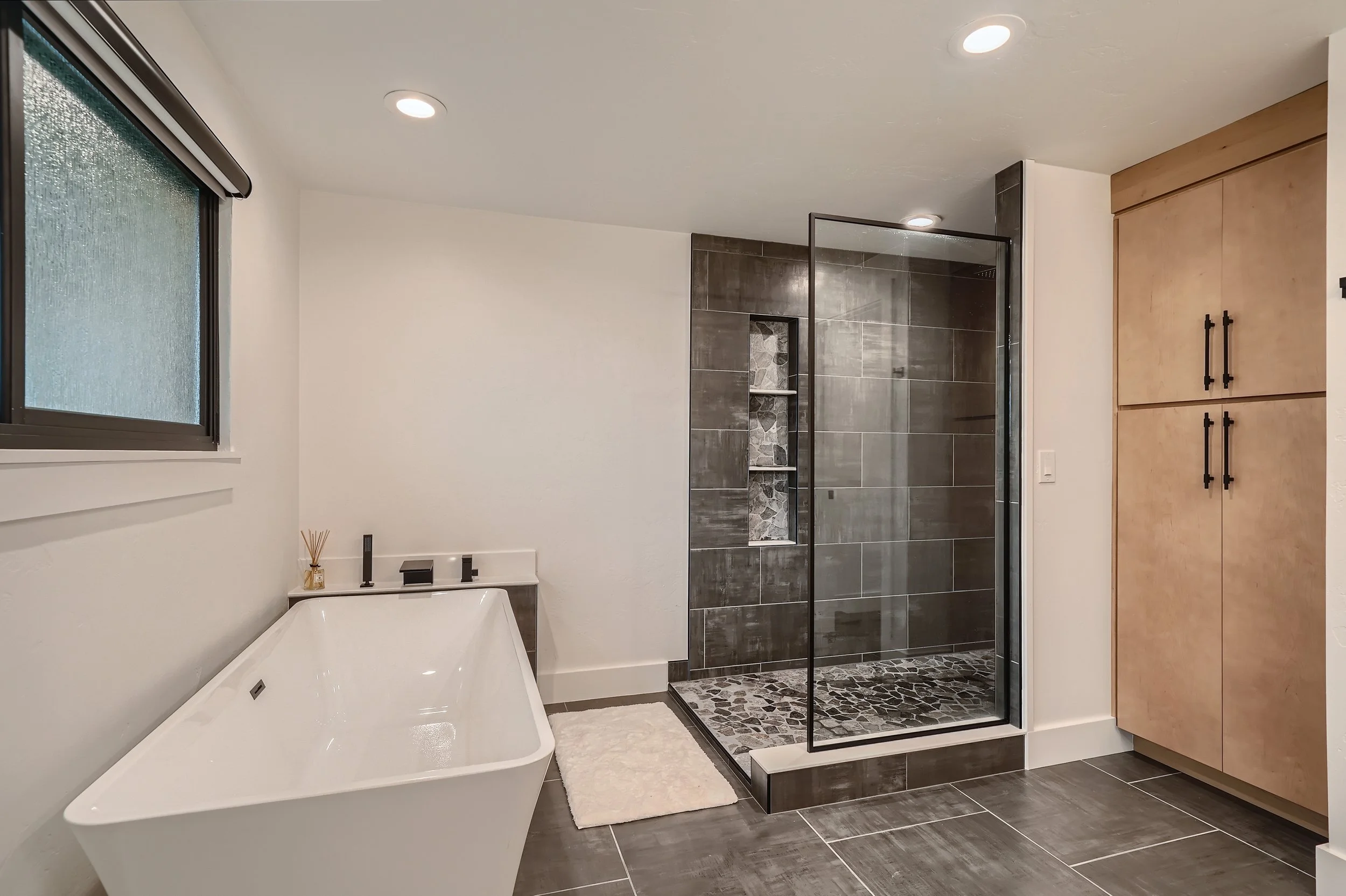Modern bathroom with freestanding bathtub, glass-enclosed shower with dark tiles and pebble floor, wooden storage cabinet, and window with frosted glass.