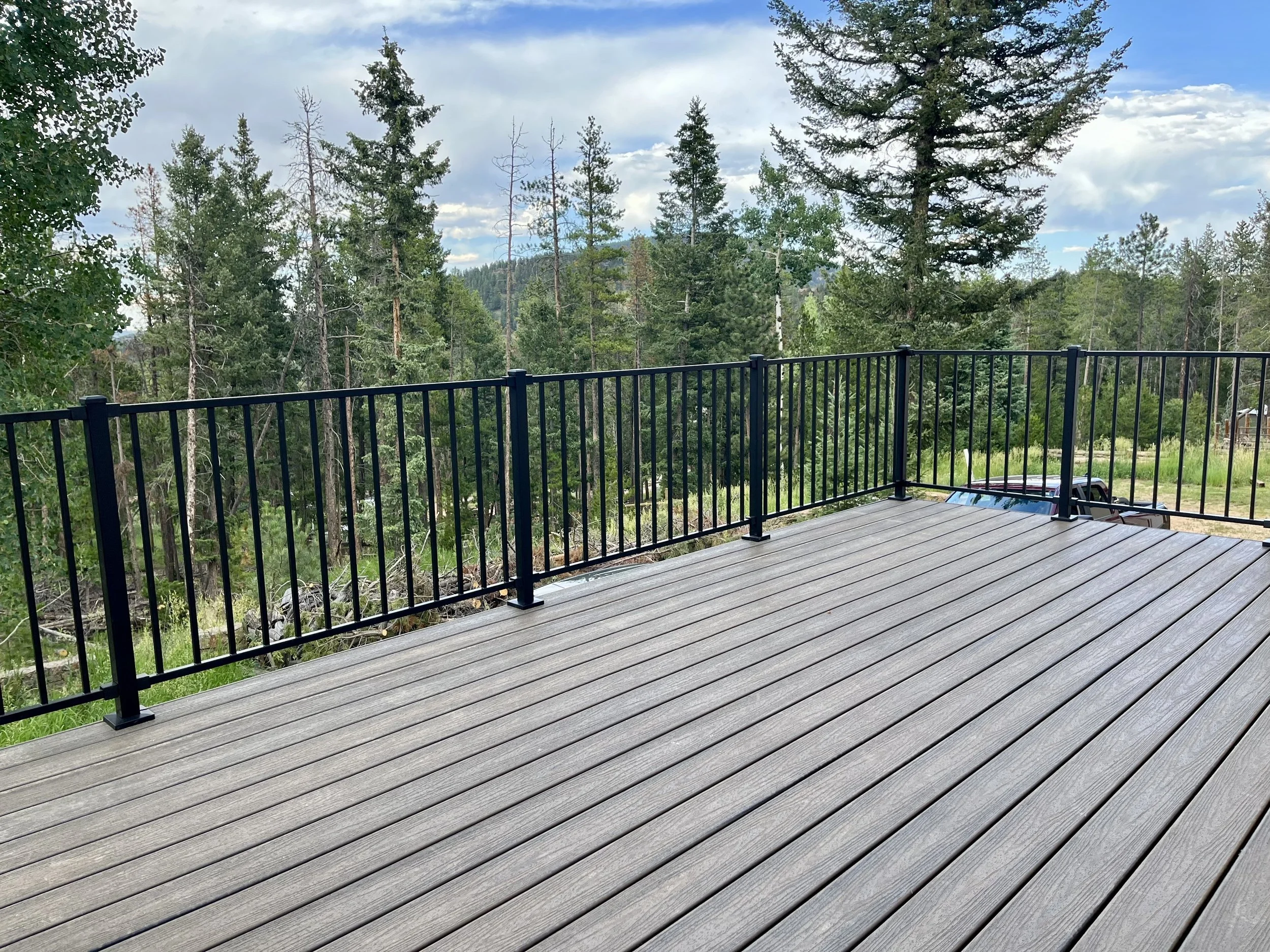 View of a wooden deck with a black metal railing overlooking a forested landscape with tall trees and a partly cloudy sky.