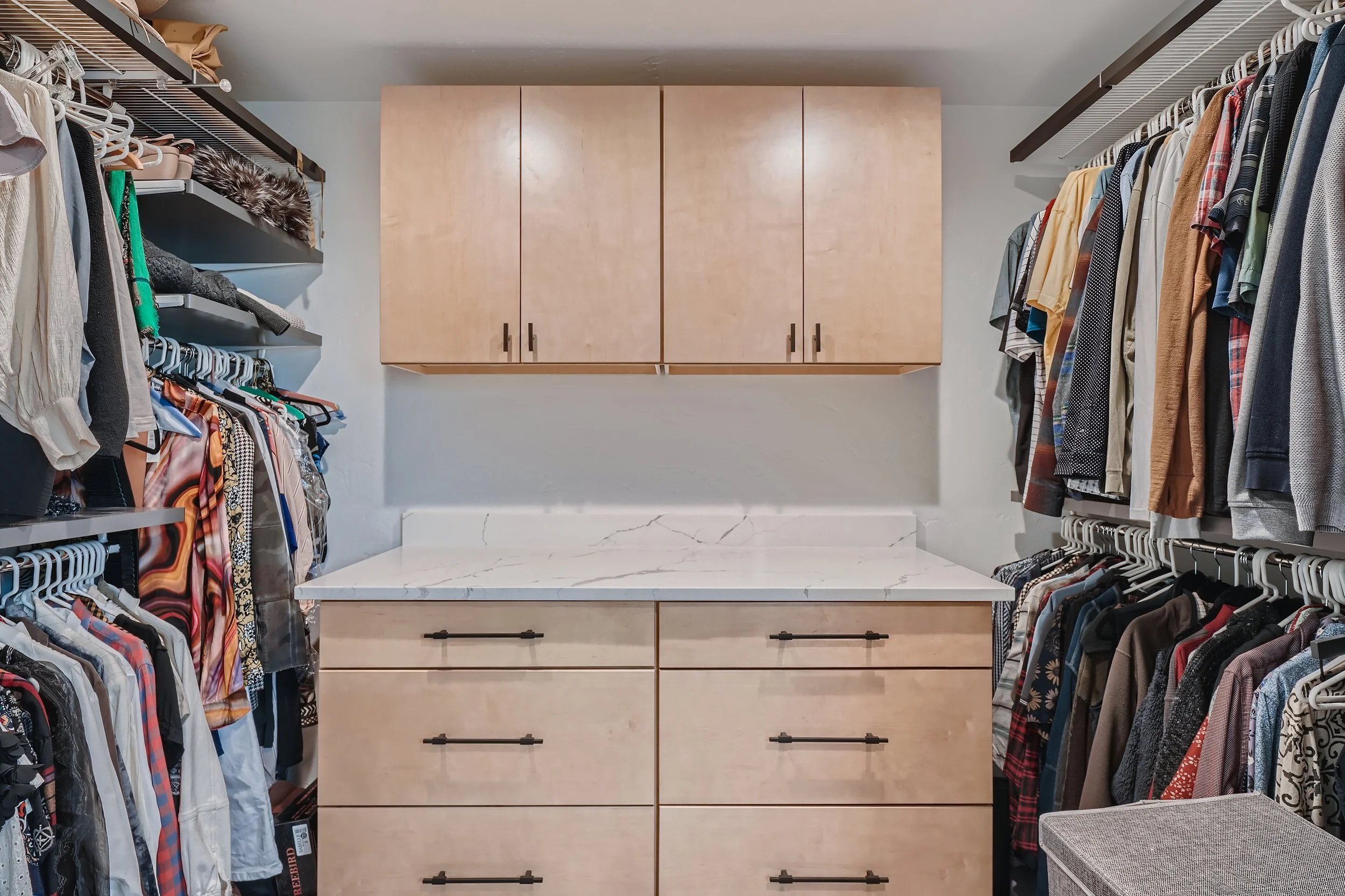 A walk-in closet with hanging clothes on both sides, a set of drawers below a marble countertop, and wall-mounted cabinets above.