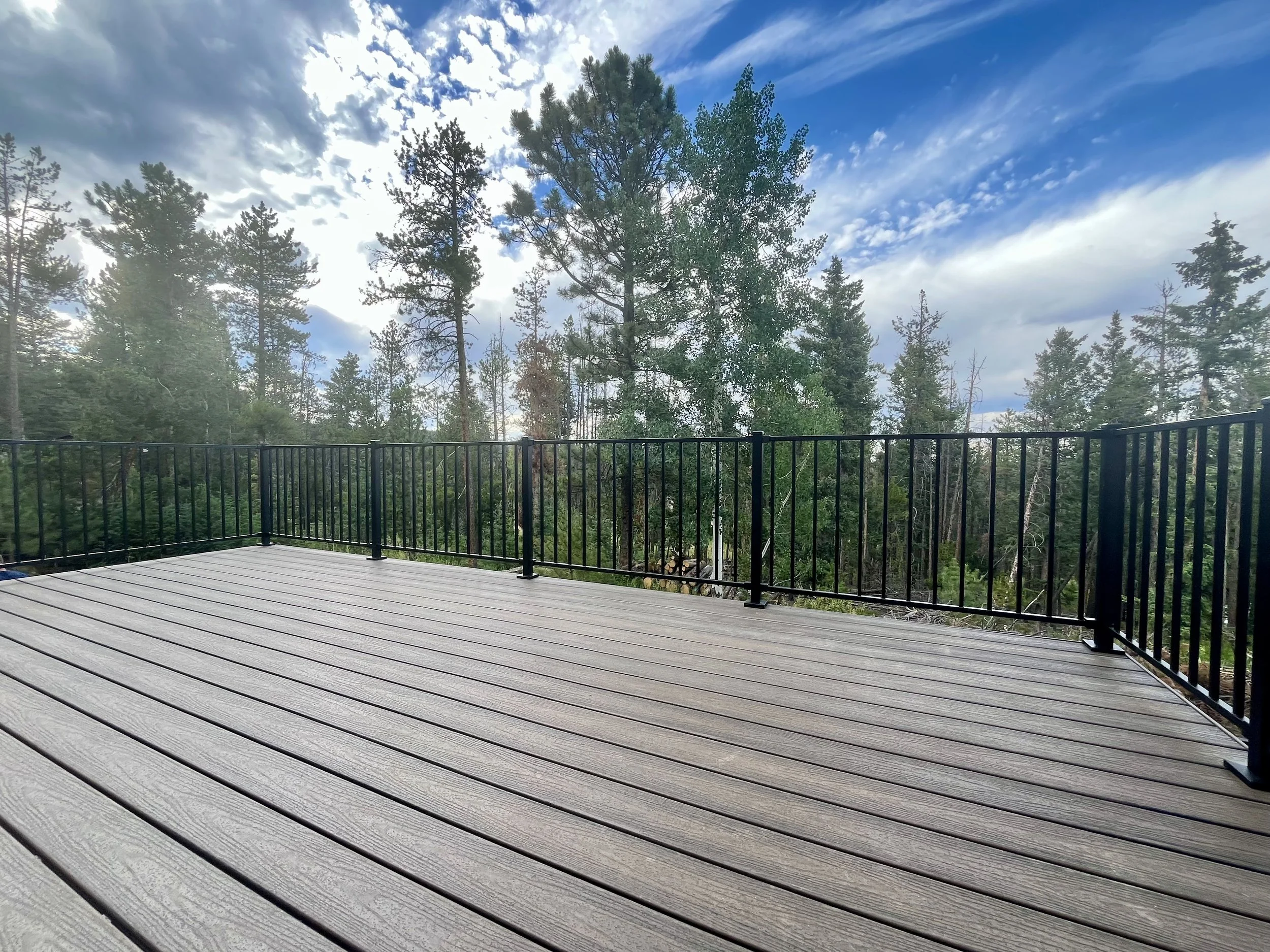 A wooden deck with black metal railing overlooking a dense forest of pine trees under a partly cloudy sky.