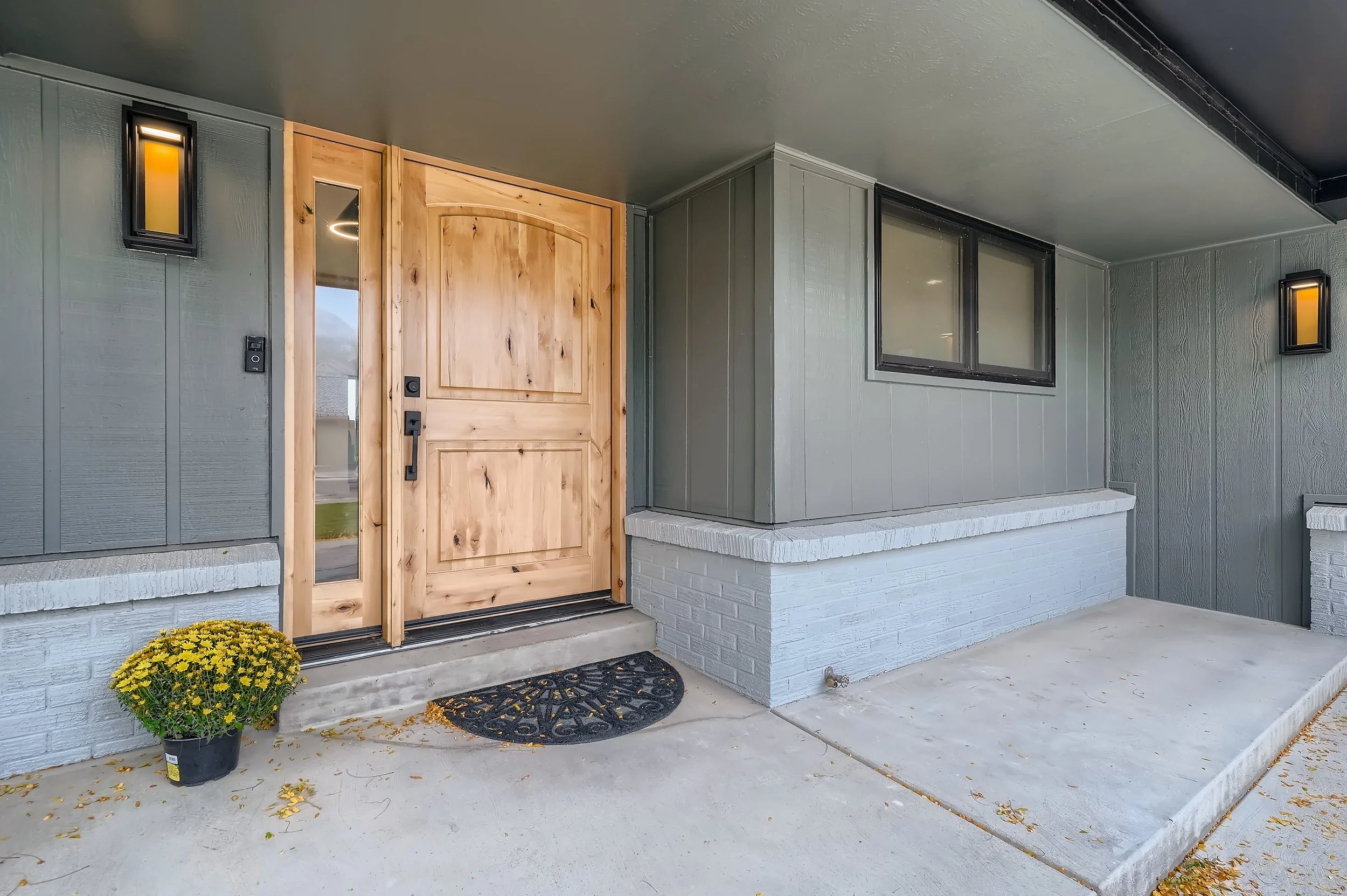 Front porch with wooden door, dark gray exterior walls, two wall-mounted lights, potted yellow flowers, and a mat.