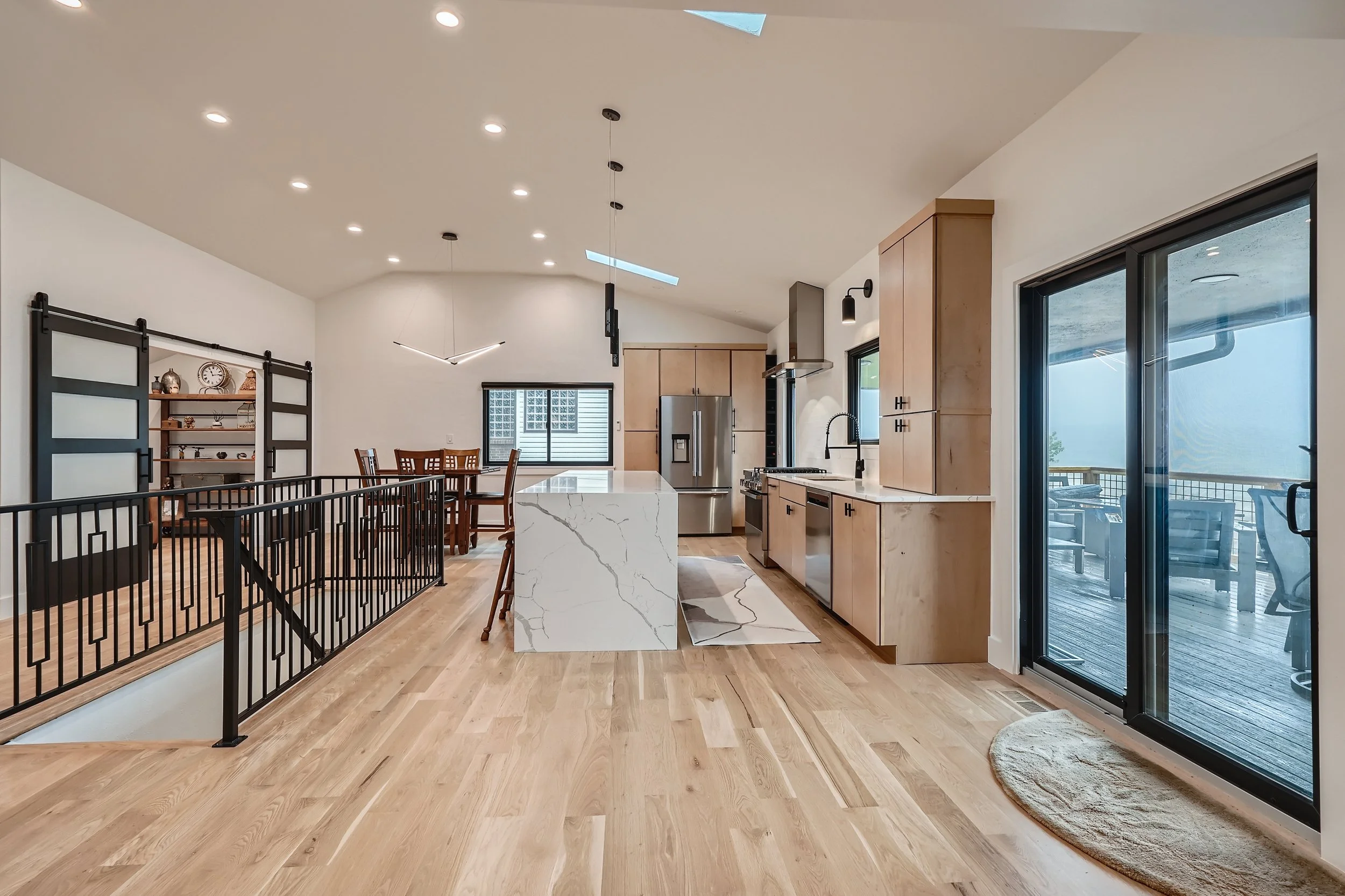 Open-concept kitchen and dining area with wooden flooring, a marble kitchen island, light wood cabinetry, stainless steel appliances, sliding glass door to balcony, and a black metal railing separating the living space from the staircase.