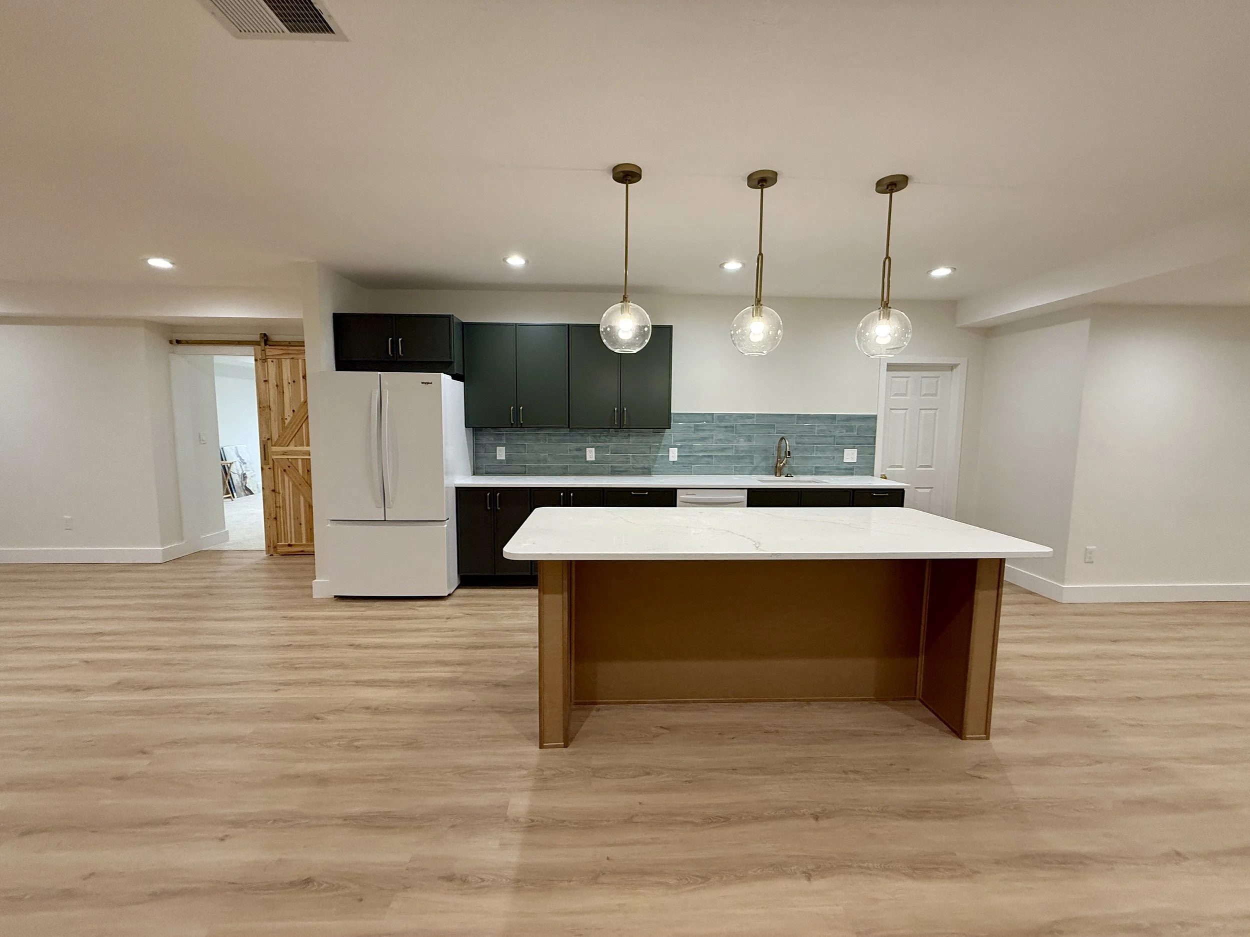Modern kitchen with dark green and black cabinets, a white refrigerator, a large central island with a white marble countertop, pendant lights, wooden flooring, and a blue tile backsplash.