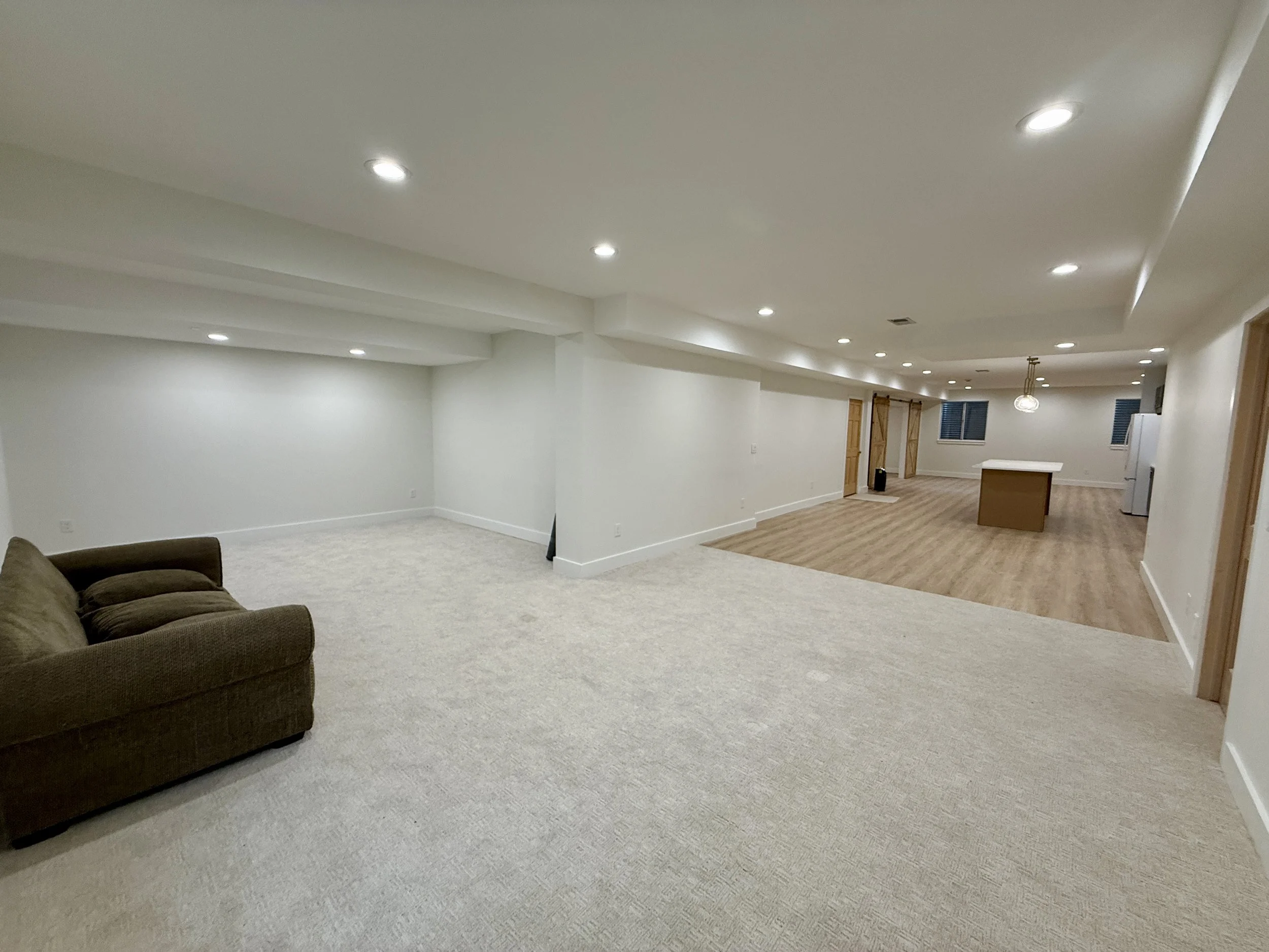 Open living area with beige carpet, brown sofa, white walls, and ceiling lights leading to a kitchen with wooden floors, a kitchen island, and white appliances.