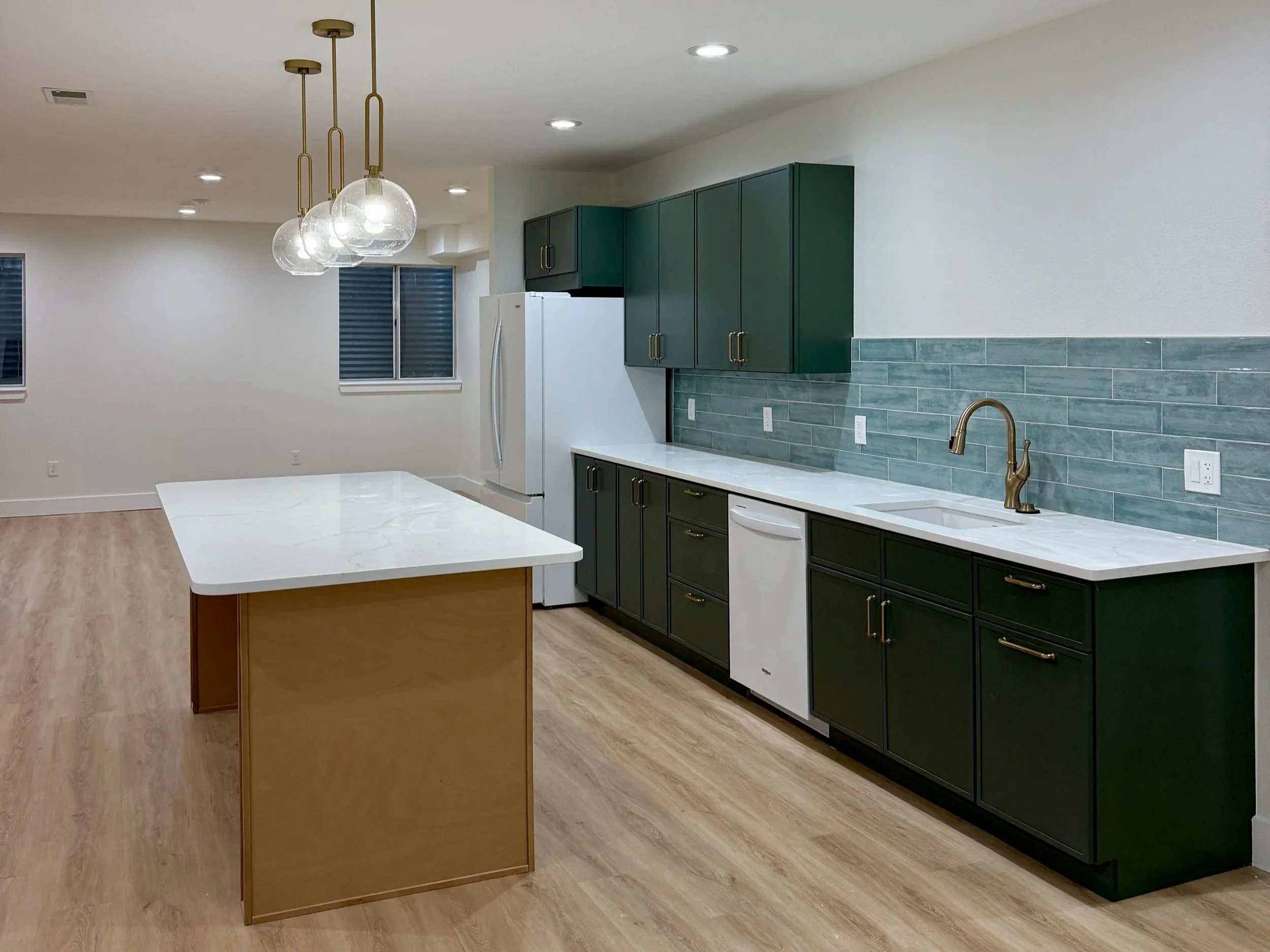 Modern kitchen with white countertops, green cabinets, a white refrigerator, and a blue tile backsplash. An island with a white marble top is in the foreground, and there are three pendant lights hanging from the ceiling.