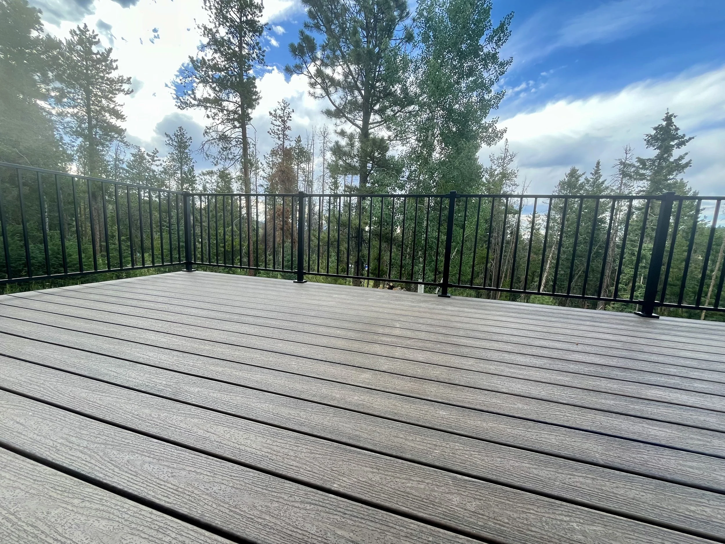 Wooden deck with black metal railing overlooking a forest of tall pine trees and a blue sky with scattered clouds.