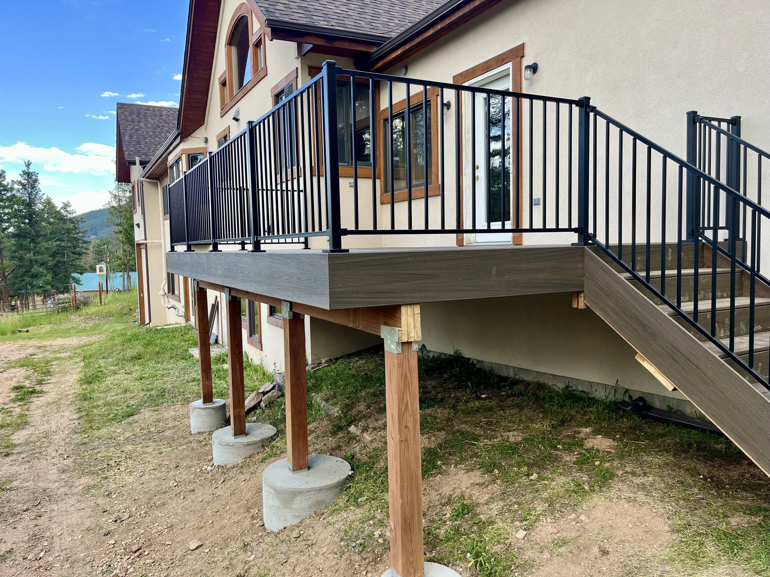 Newly built house with a wooden deck and black metal railing, elevated on wooden supports and concrete footings, on a grassy lot with mountains and trees in the background under a blue sky.