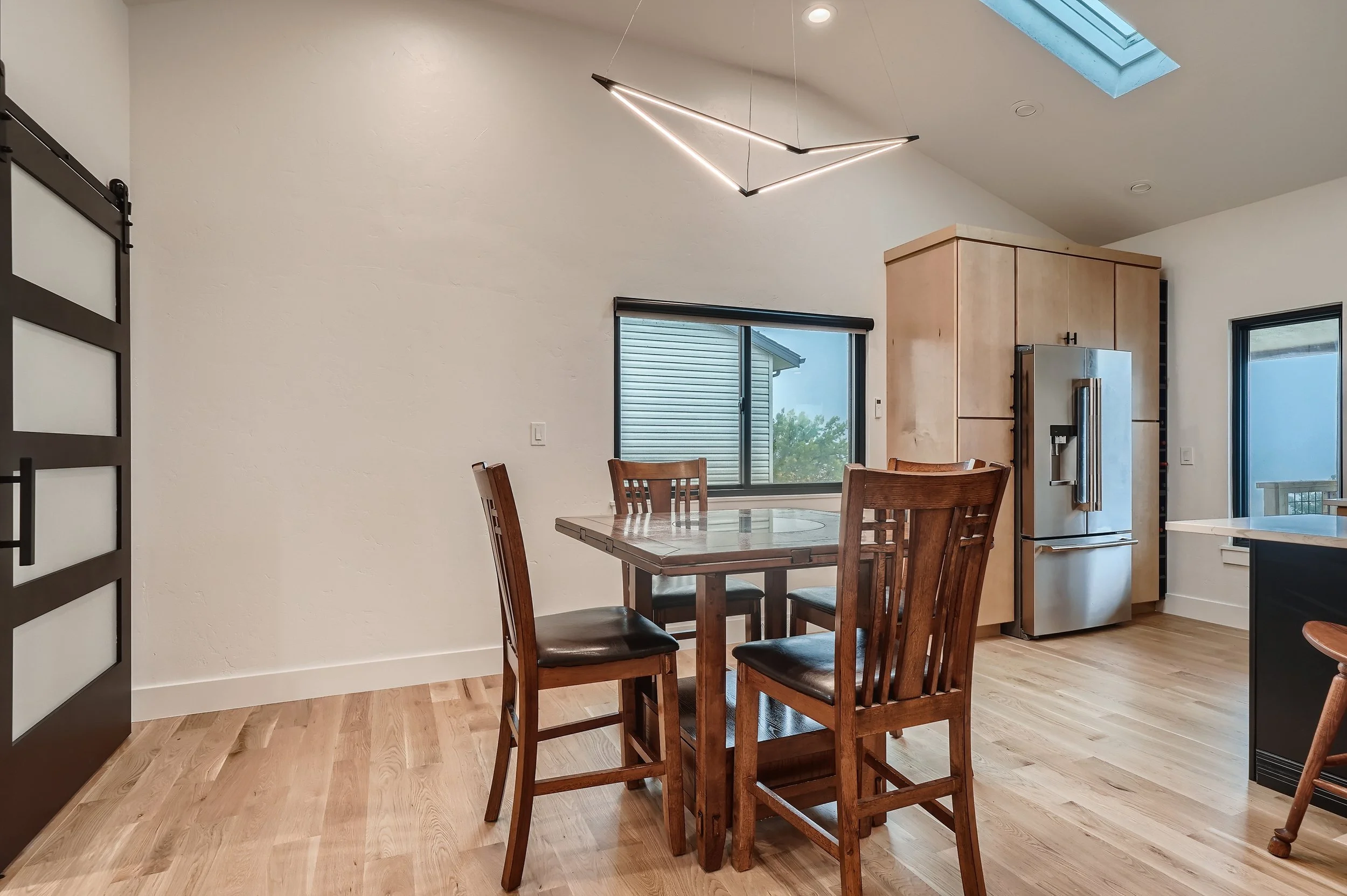 Kitchen with wooden dining table and four chairs, wooden cabinetry, stainless steel refrigerator, window, sliding glass door, skylight, wooden flooring, modern geometric ceiling light.