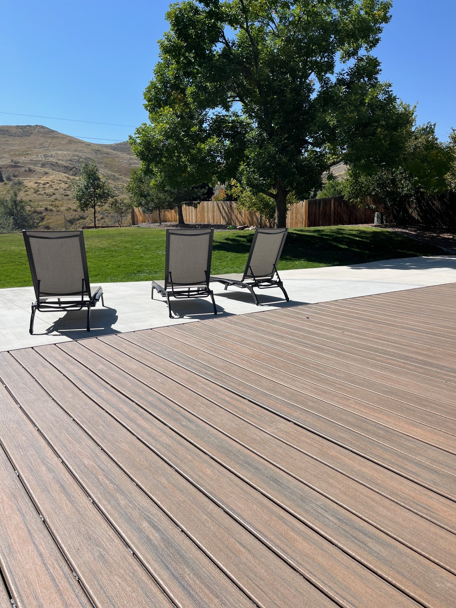 Three lounge chairs on a patio next to a grassy yard with a large tree and hills in the background.