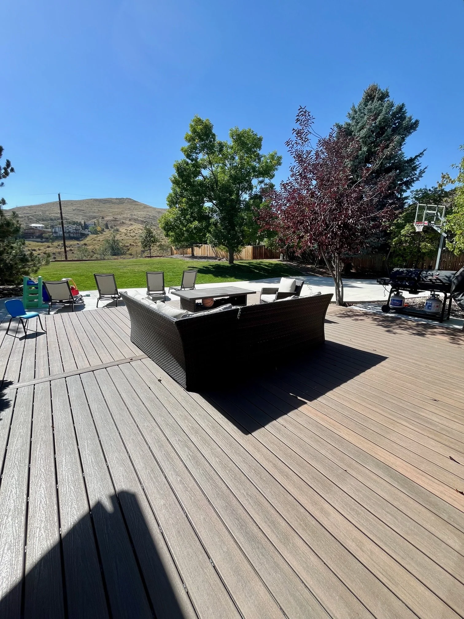 A backyard deck with outdoor furniture, including a wicker couch, a coffee table, and several chairs. The yard has a grassy area, trees, a fence, and a mountain in the background. There is also a small playhouse, a basketball hoop, and a grill.