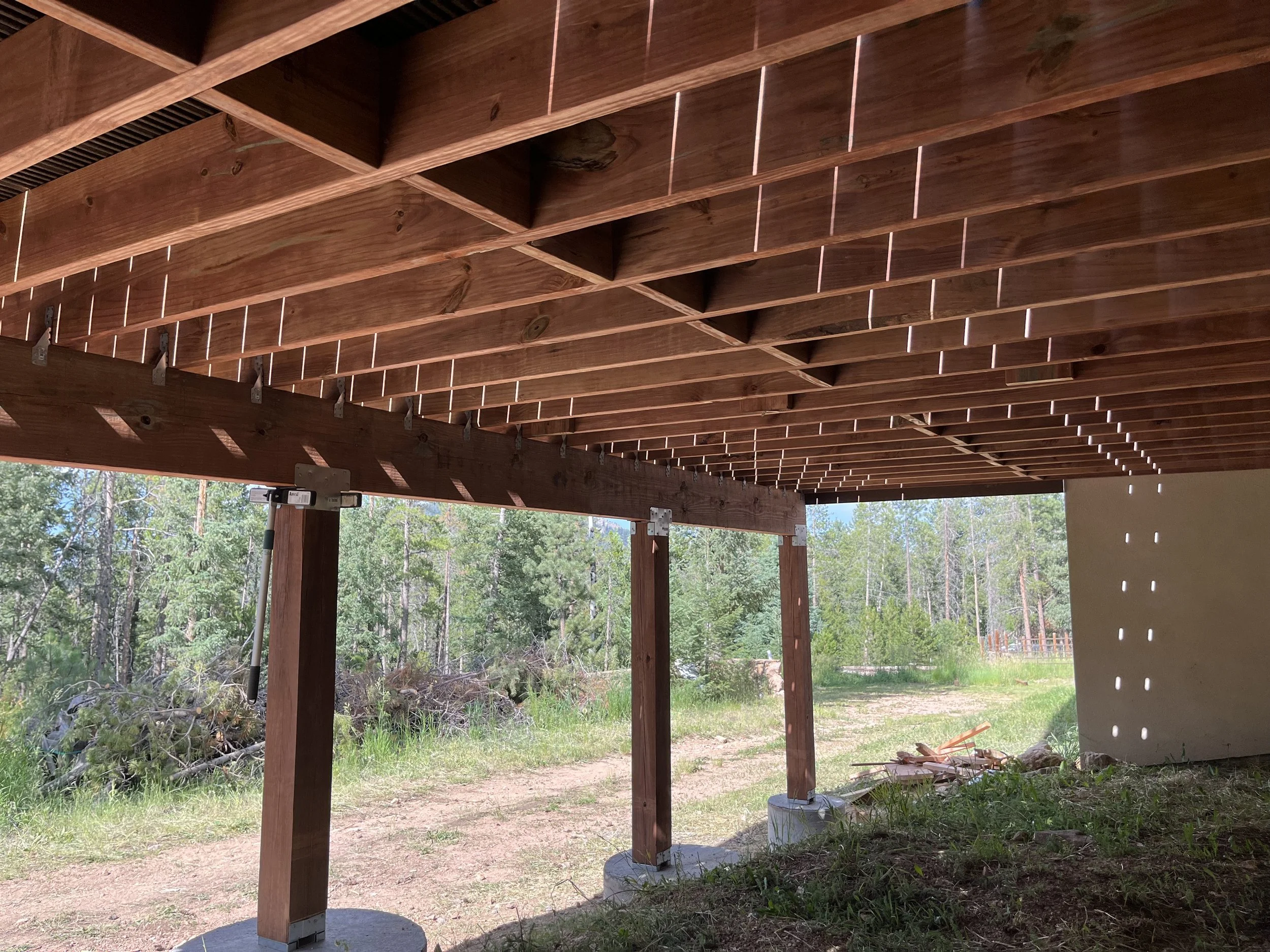 Underneath a wooden deck or porch with sunlight filtering through gaps in the wooden planks, supported by wooden posts on concrete footings, with a natural outdoor background of trees and grass.