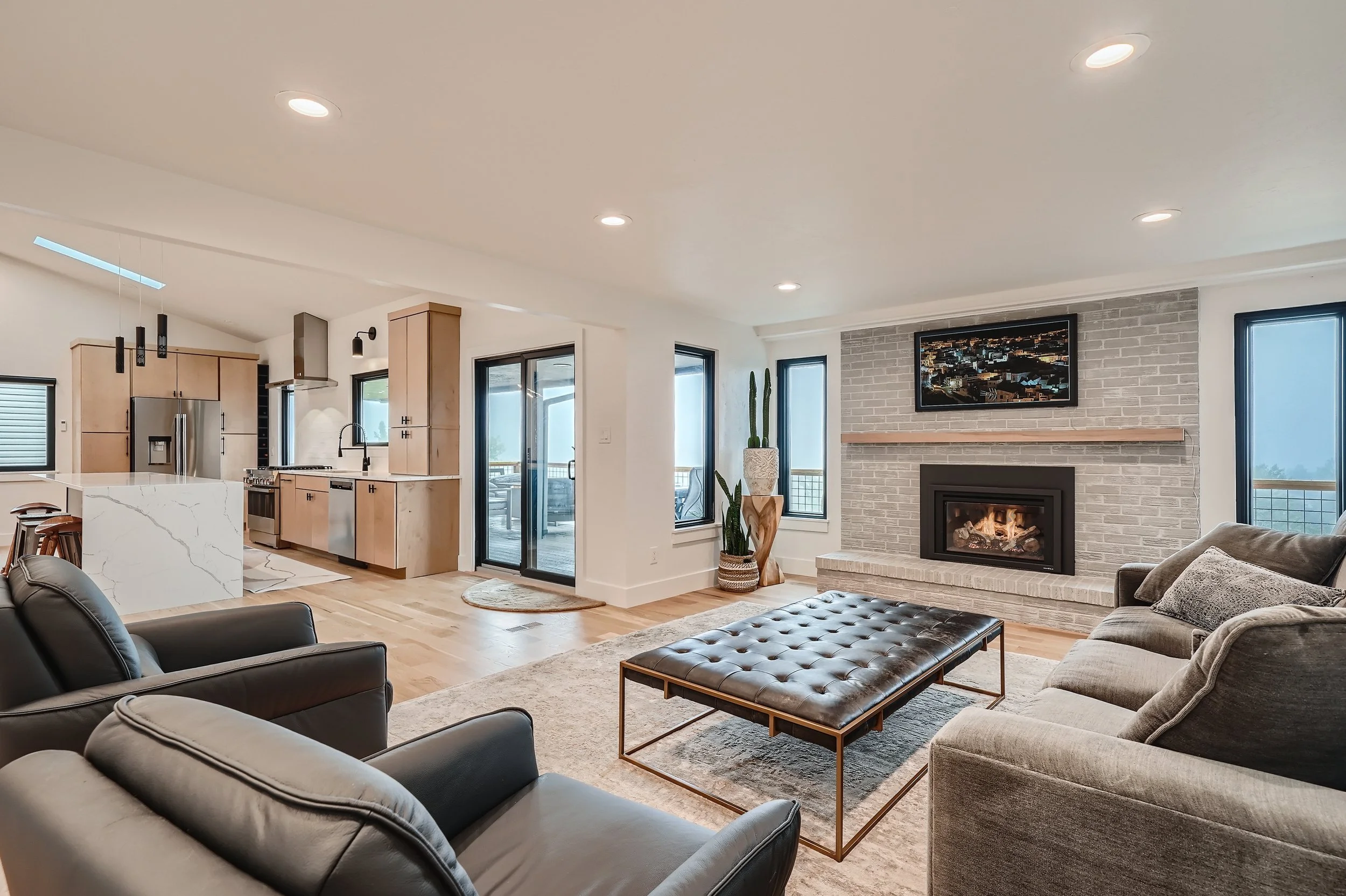 Open-concept living room with a fireplace, gray couches, a black leather ottoman, and a wall-mounted TV, adjacent to a kitchen with light wood cabinets and marble island, with glass doors leading to an outdoor patio.