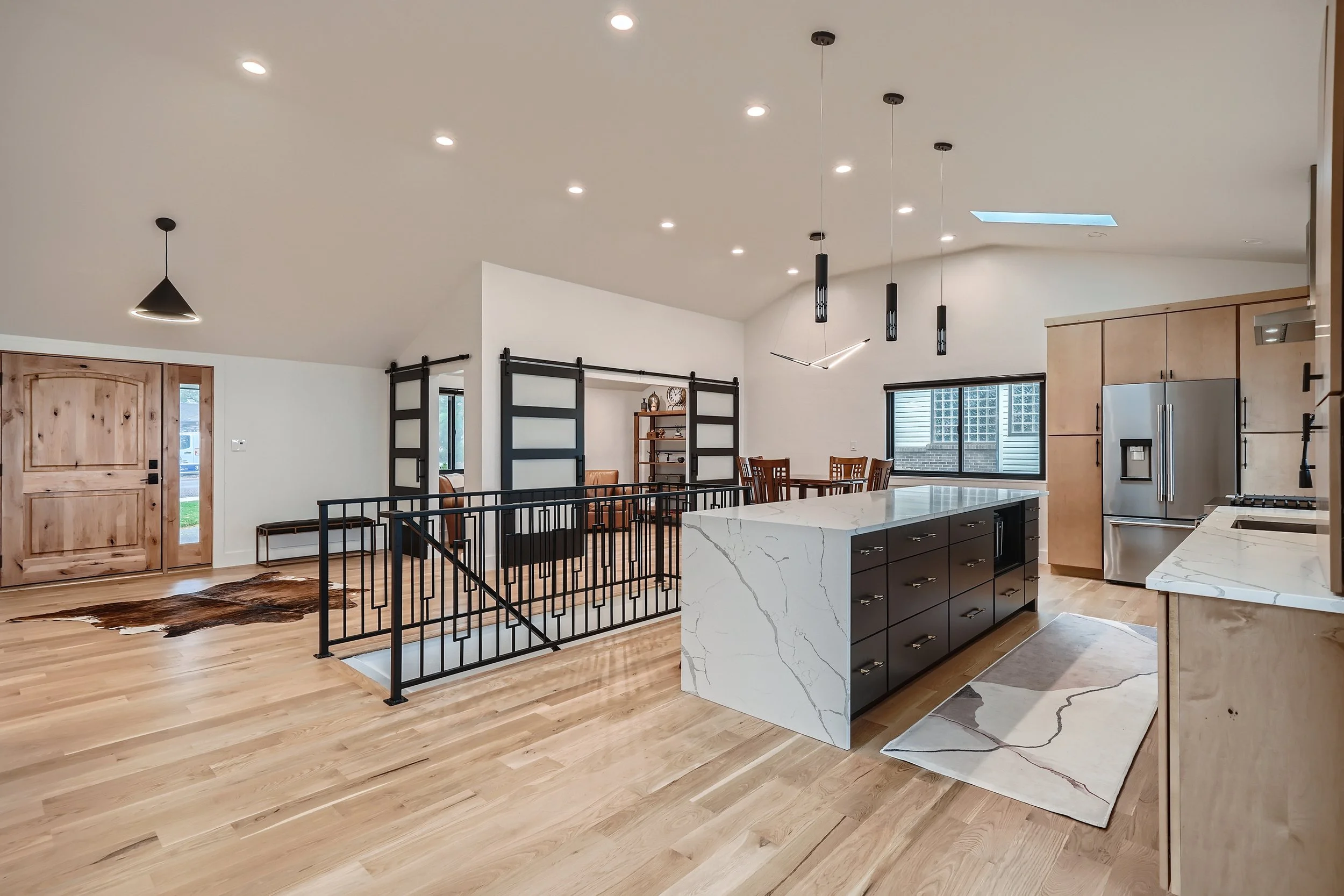 Open-concept kitchen and living area with wooden flooring, a marble kitchen island, stainless steel appliances, and modern lighting fixtures.