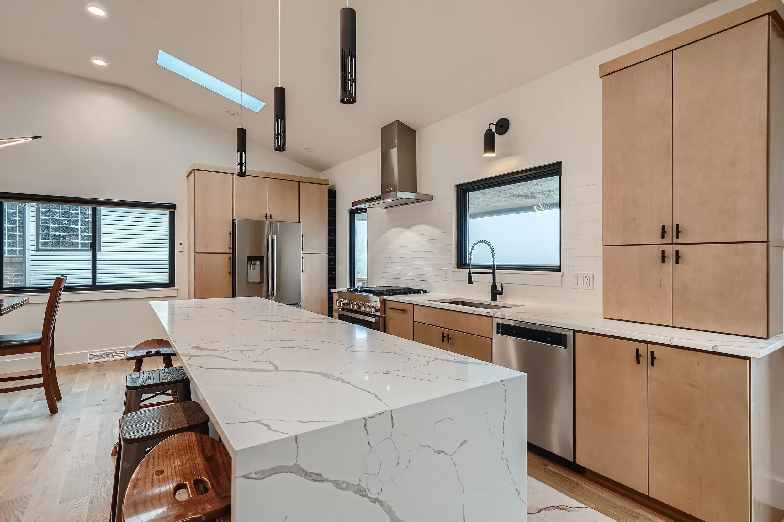 Modern kitchen with light wood cabinets, white marble countertops, stainless steel appliances, black window frames, pendant and wall lighting, a skylight, and a large island with bar stools.