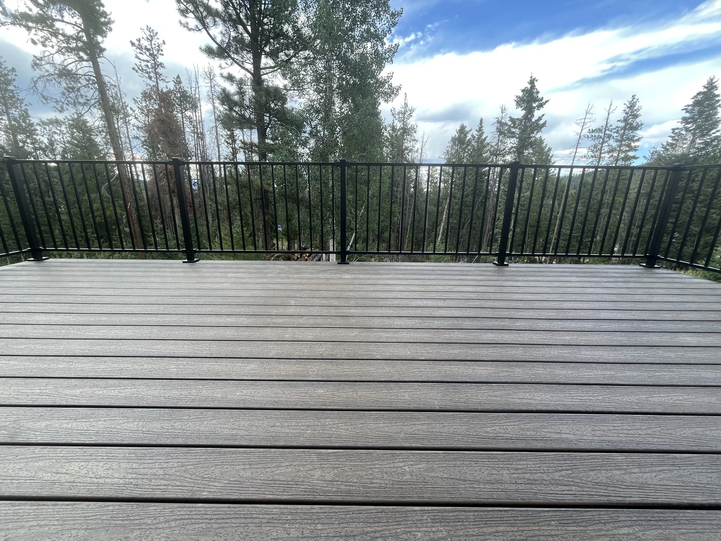 Wooden deck with black metal railing overlooking a forest of tall pine trees under a partly cloudy sky.