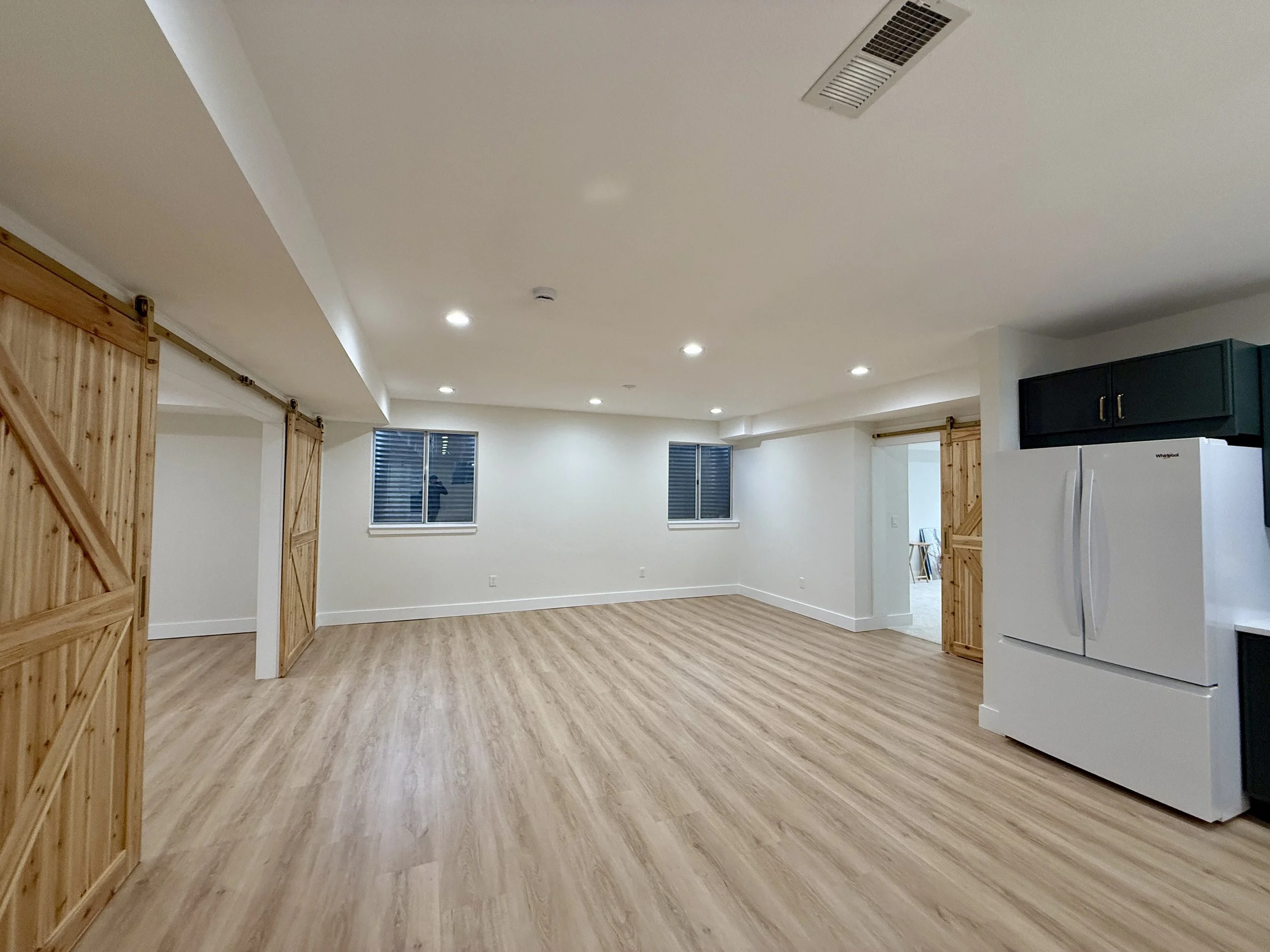 Empty basement with wood-look flooring, white walls, small windows, wood sliding barn doors, and a refrigerator.