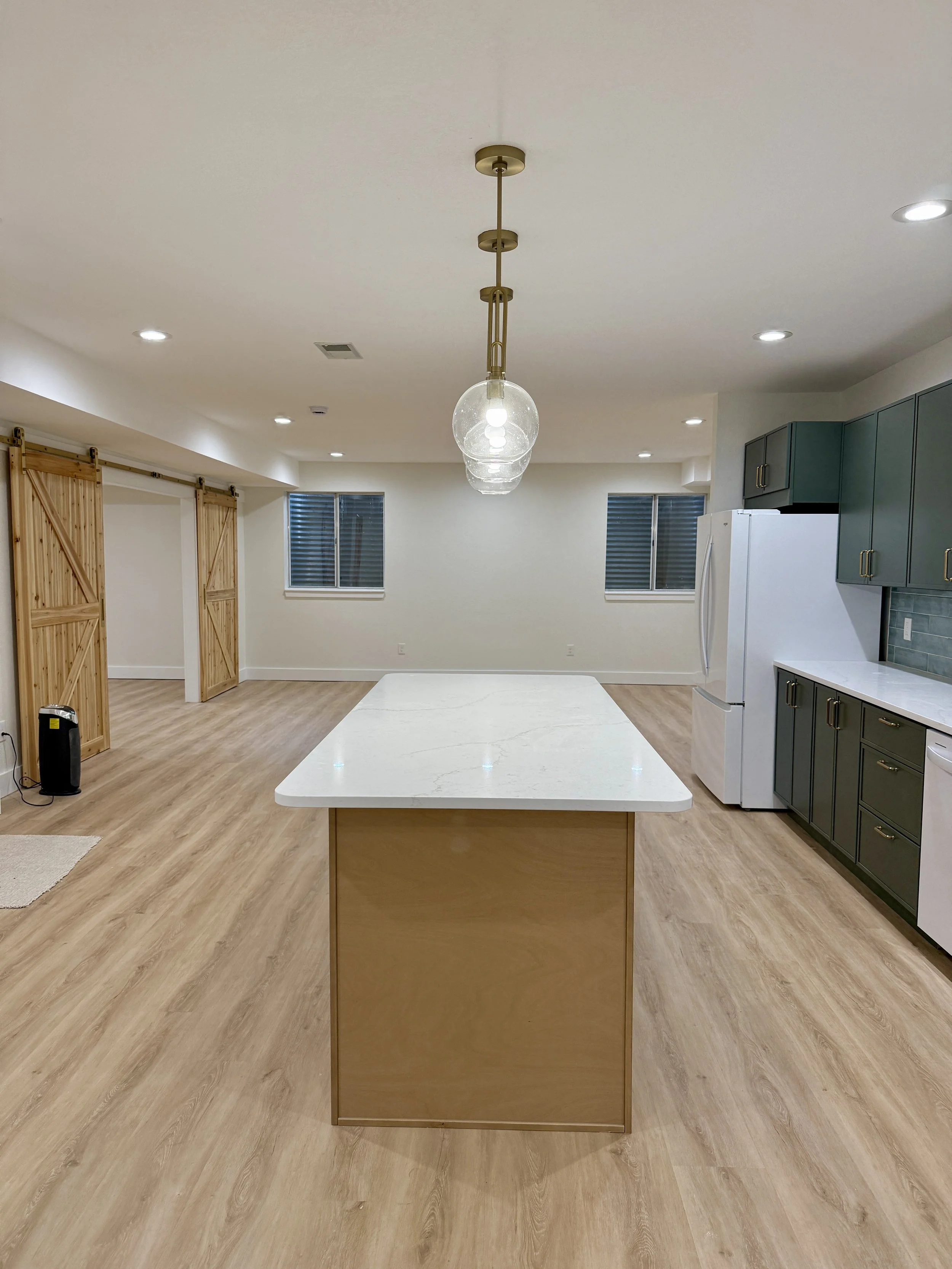 Modern kitchen with a large white island, green cabinets, white refrigerator, three windows, and wooden sliding barn doors.