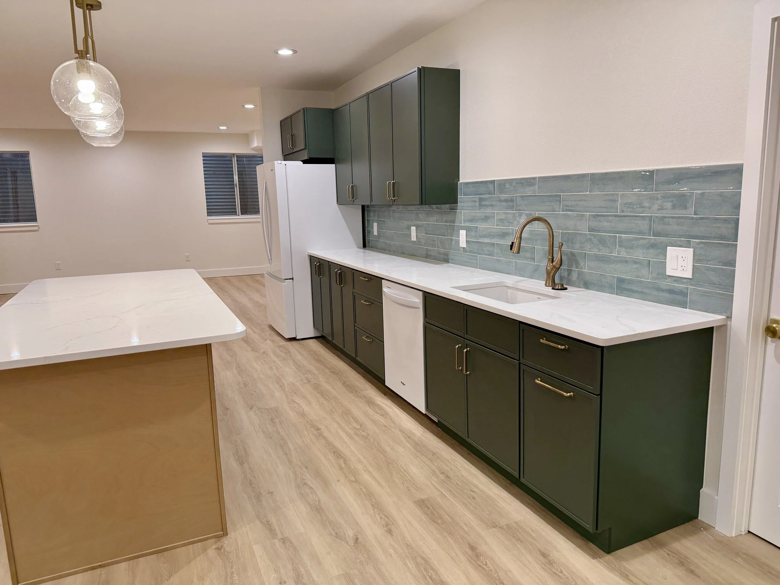 Kitchen with green cabinets, white marble countertop, blue tiled backsplash, stainless steel faucet, white refrigerator, dishwasher, light wood flooring, and pendant lights.