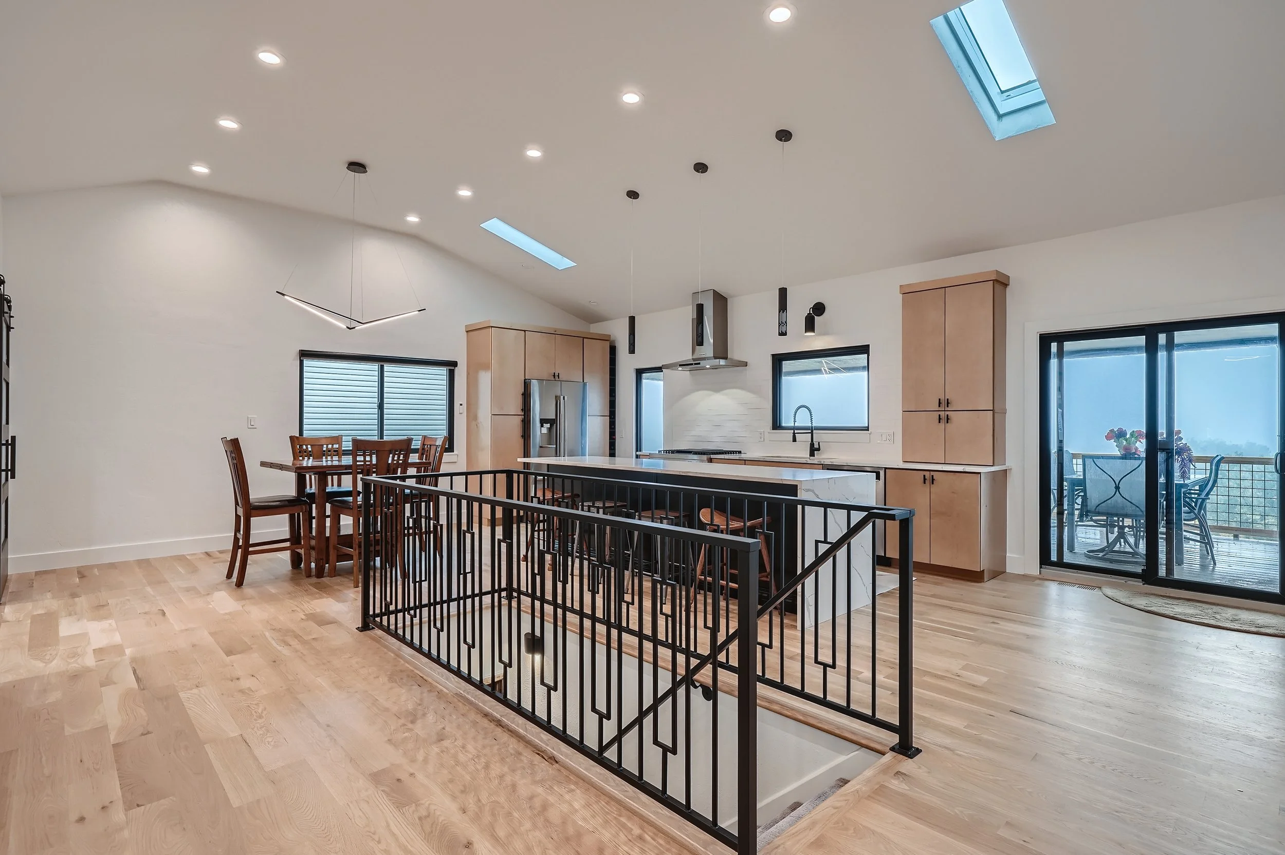Open-concept kitchen and dining area with natural light, light wood floors, pendant lights, skylights, sliding glass doors, and a staircase with a black metal railing.