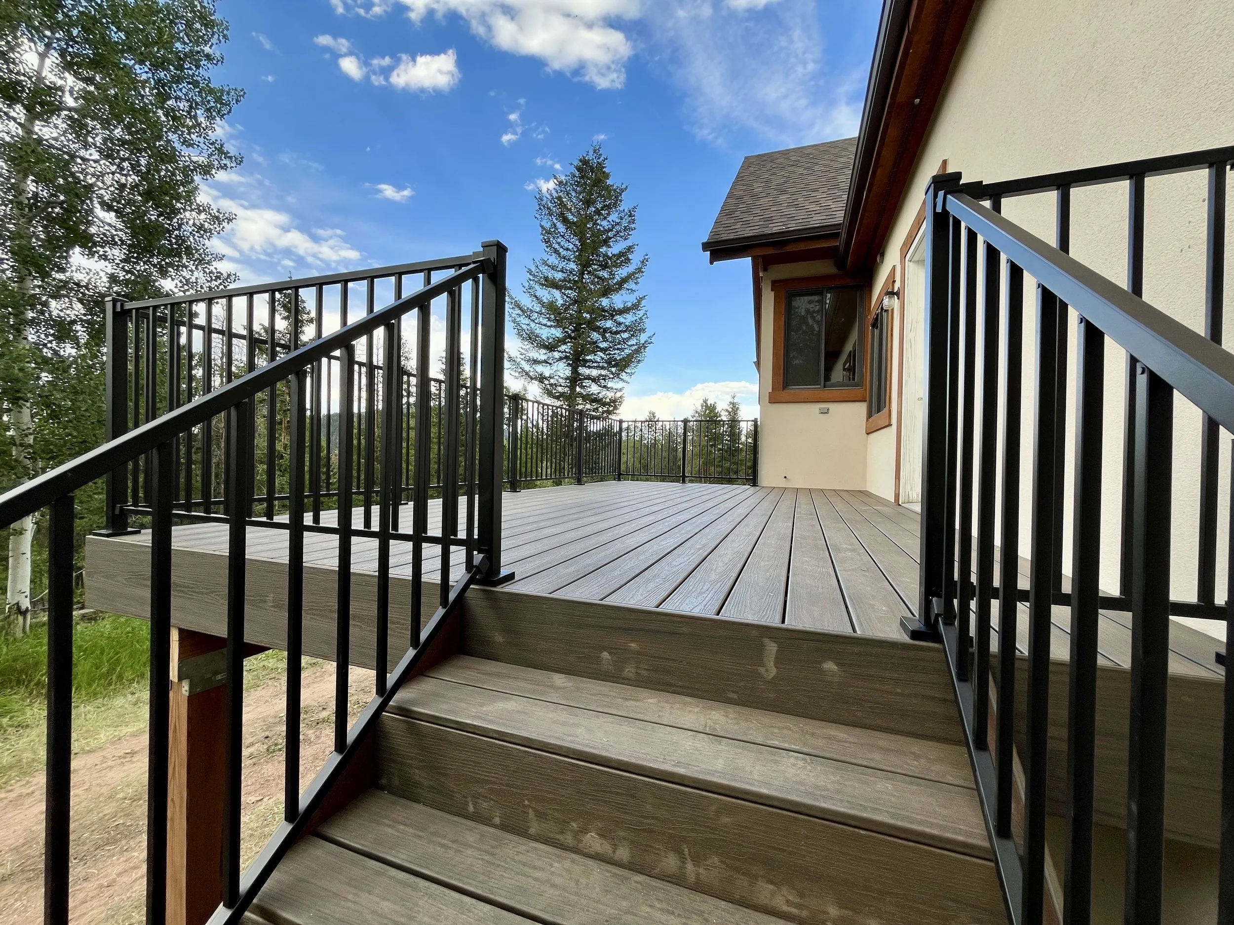 Wooden deck with black metal railing outside a house, surrounded by trees and a blue sky with some clouds.