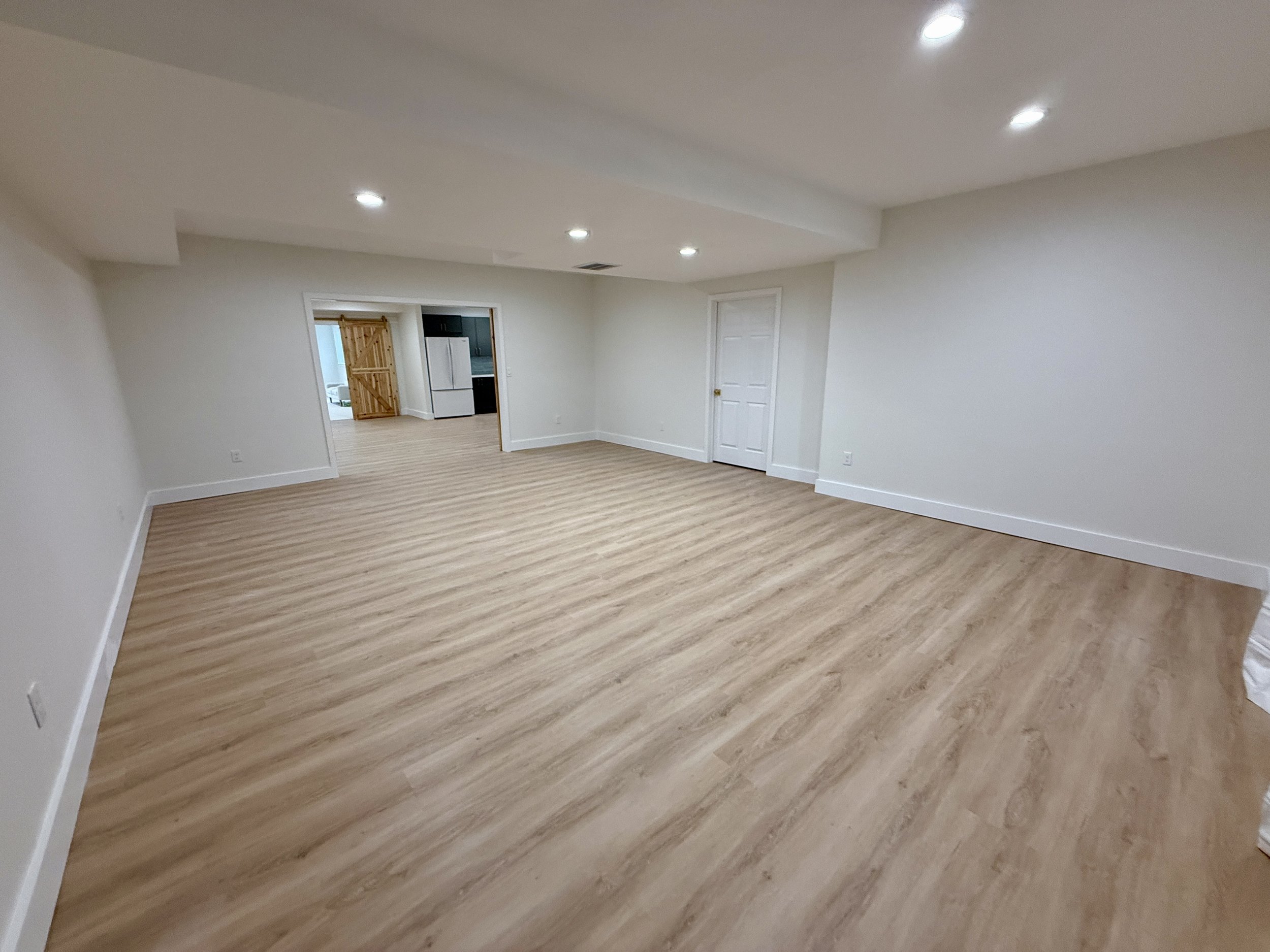 Empty room with white walls, light wood flooring, and recessed ceiling lights, leading to a kitchen area in the background.