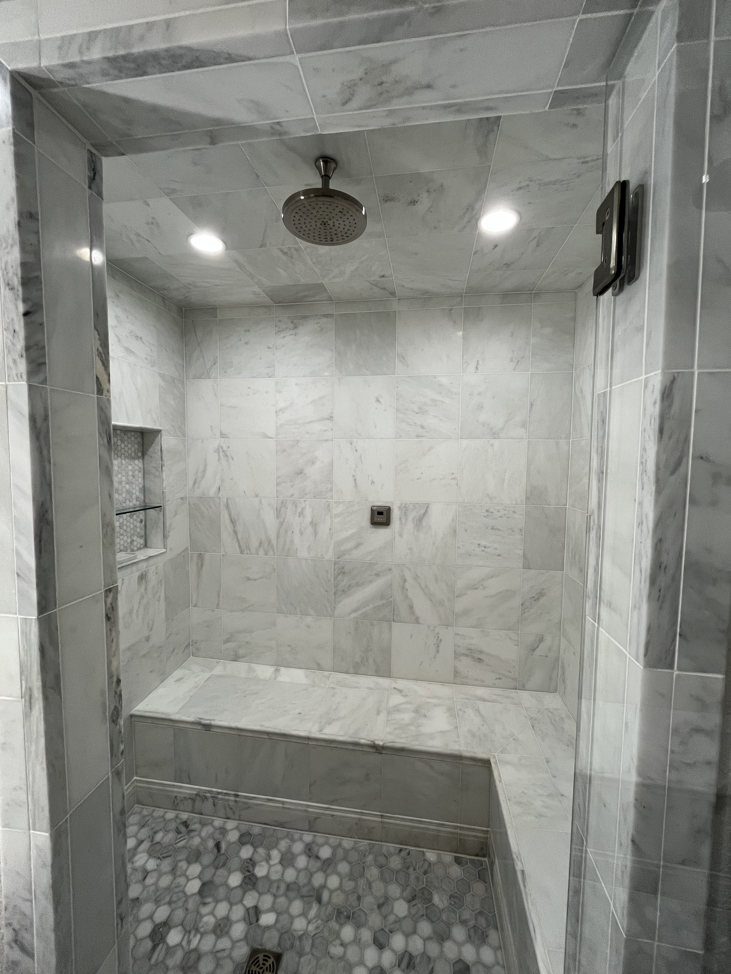 Shower stall with marble tiles, a rain shower head on the ceiling, recessed ceiling lights, a built-in niche with shelves, and a bench, with a gray hexagonal tile floor.