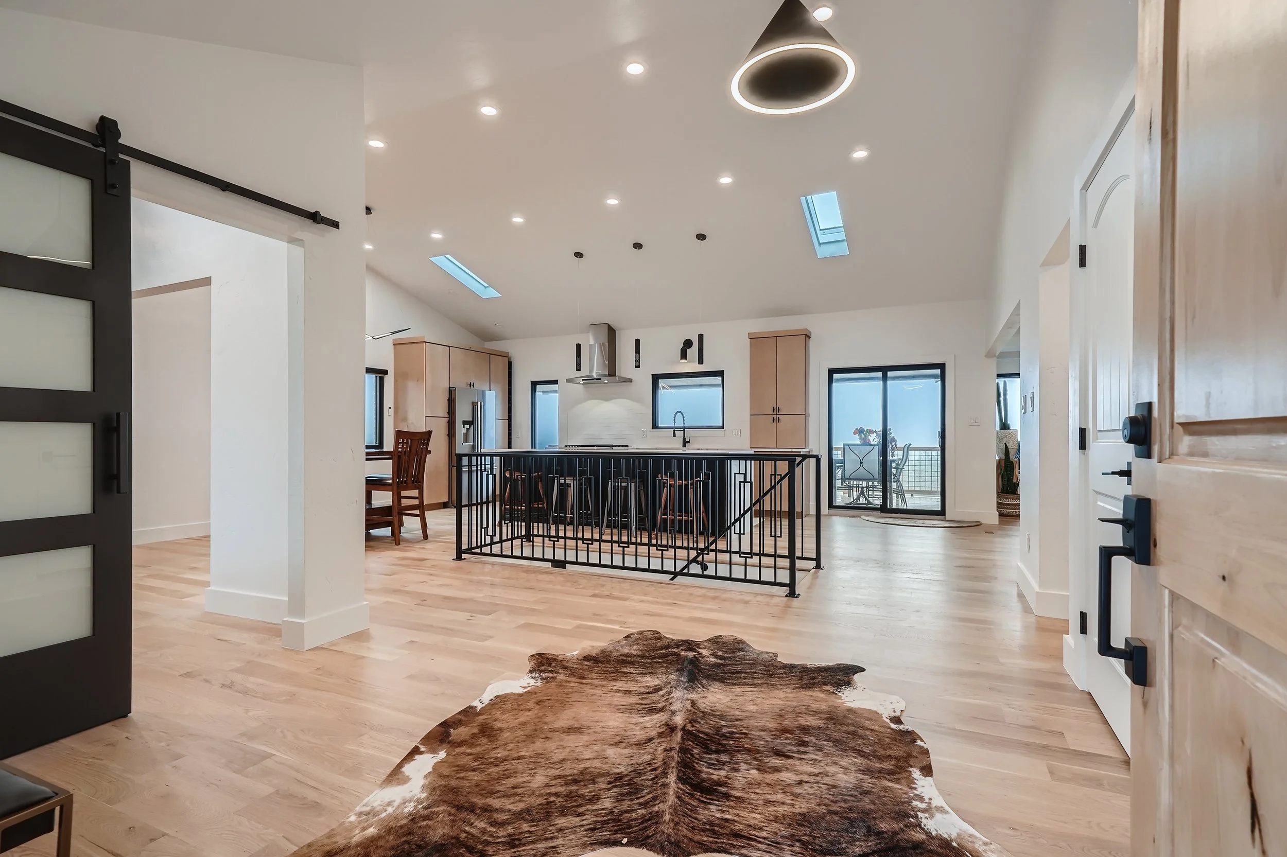 Open-concept kitchen and living space with light wood flooring, modern kitchen appliances, and a cowhide rug in the foreground. Natural light streams through skylights and large sliding glass doors, leading to an outdoor patio.