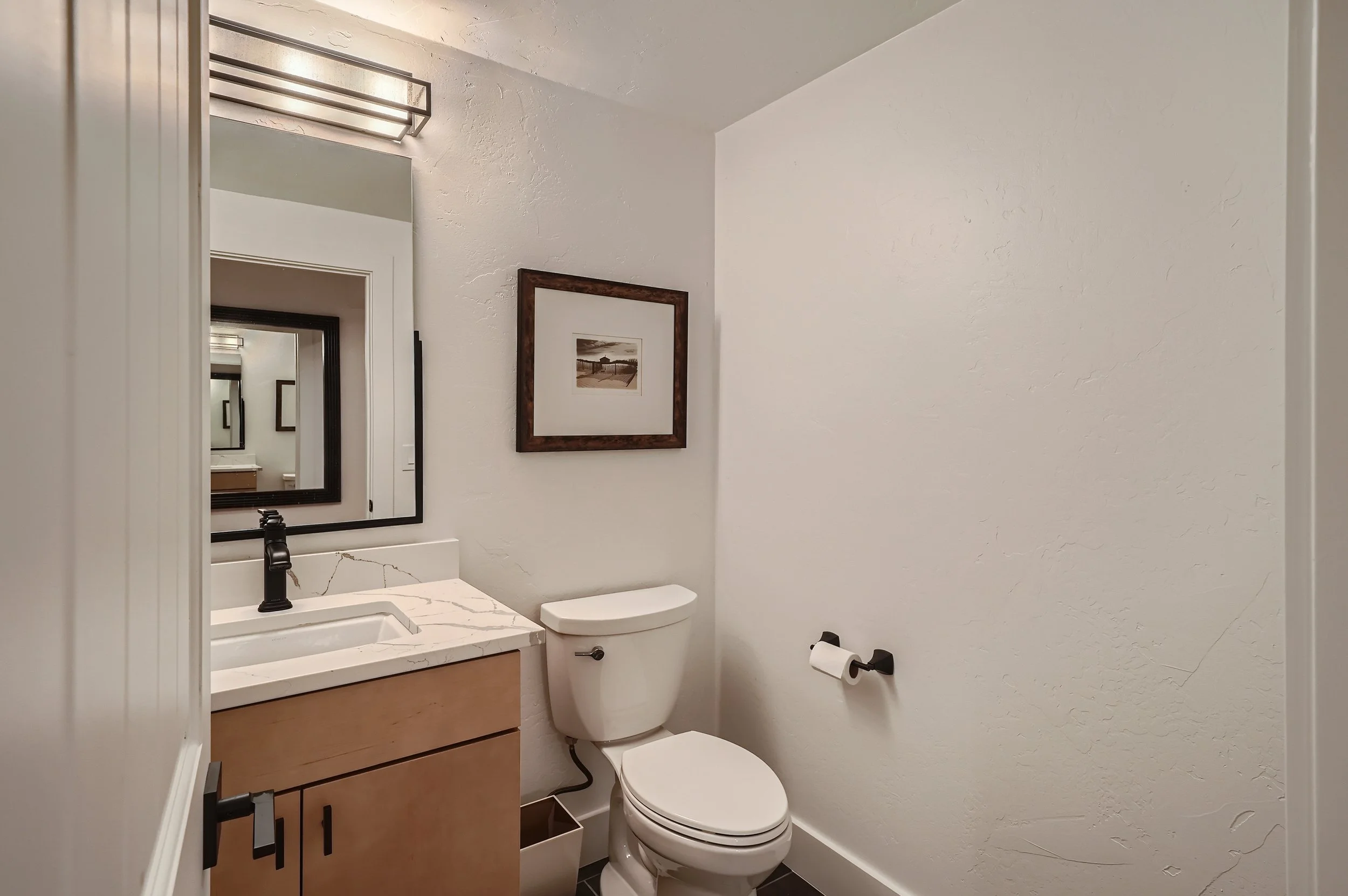 Small bathroom with white walls, a toilet, and a wooden vanity with a marble countertop and black faucet. A framed picture hangs on the wall above the toilet, and a mirror is reflected in the small bathroom mirror. A black toilet paper holder is moun