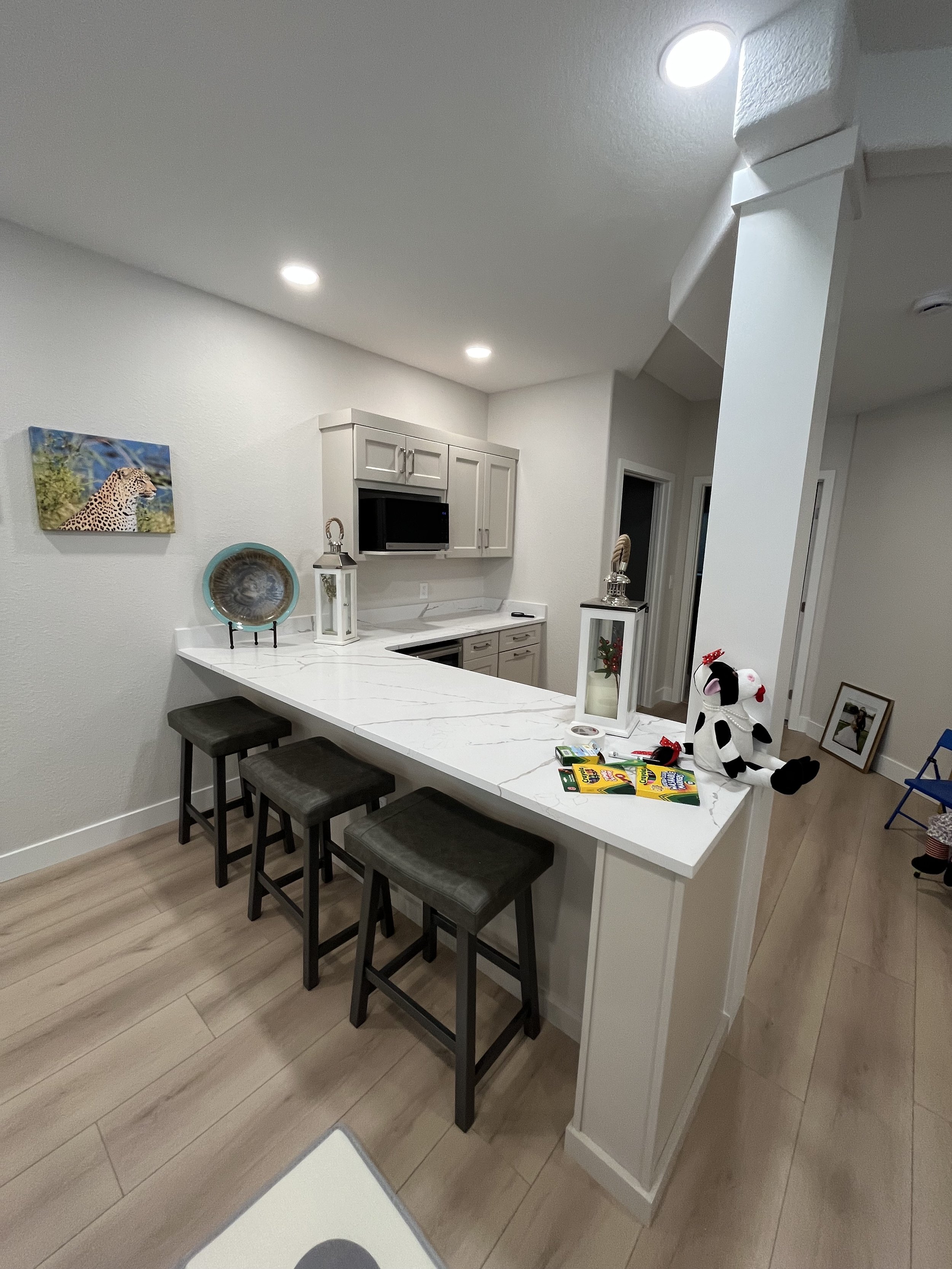 A kitchen with white walls, light wood flooring, a white marble breakfast bar with three dark green bar stools, a white upper cabinet with a microwave, and front door. Decor includes a painting of a jaguar, a blue tray, a lantern, a plush dog toy, an