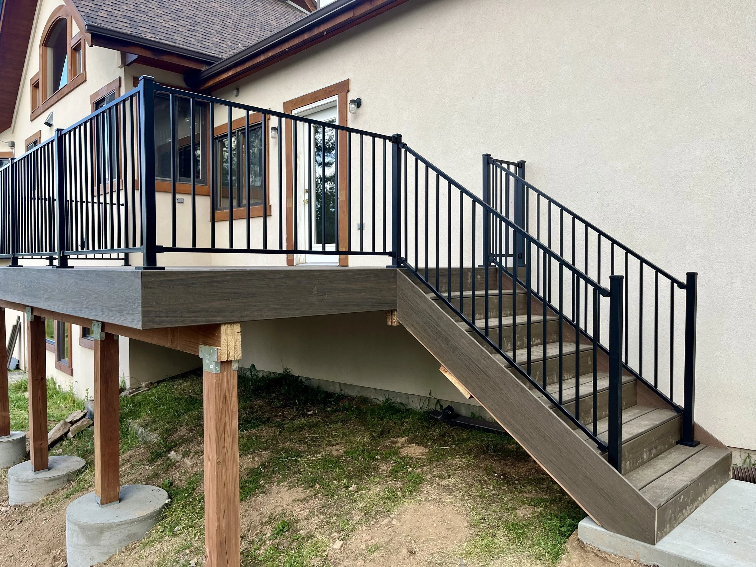Newly installed outdoor deck with black metal railing and stairs leading down to yard, attached to beige house with wooden window frames and a glass door.