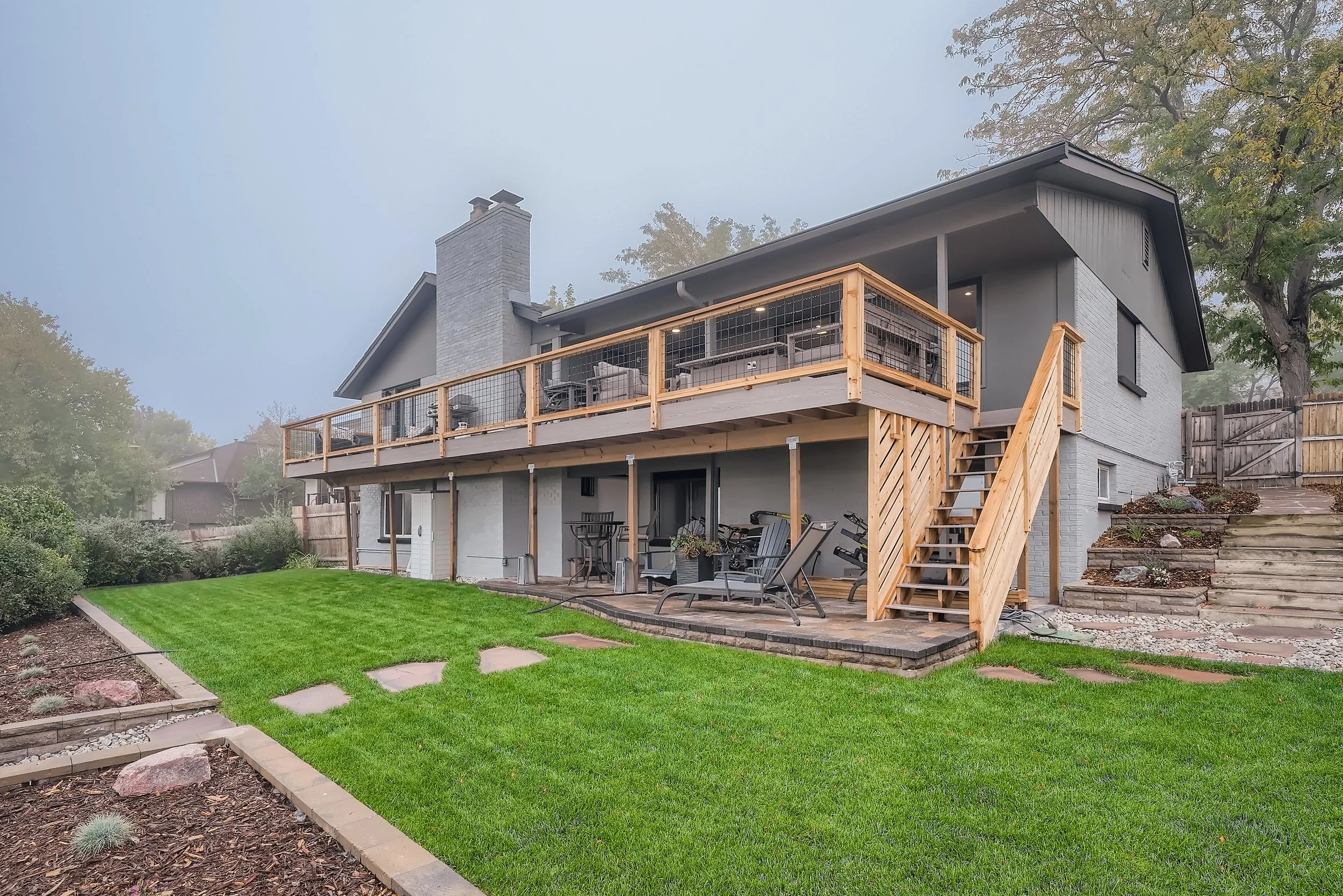 Backyard of a house with a well-maintained lawn, a wooden deck with outdoor furniture, exercise equipment, and a staircase leading to the upper porch. The house has gray siding, a chimney, and is surrounded by trees and a wooden fence.