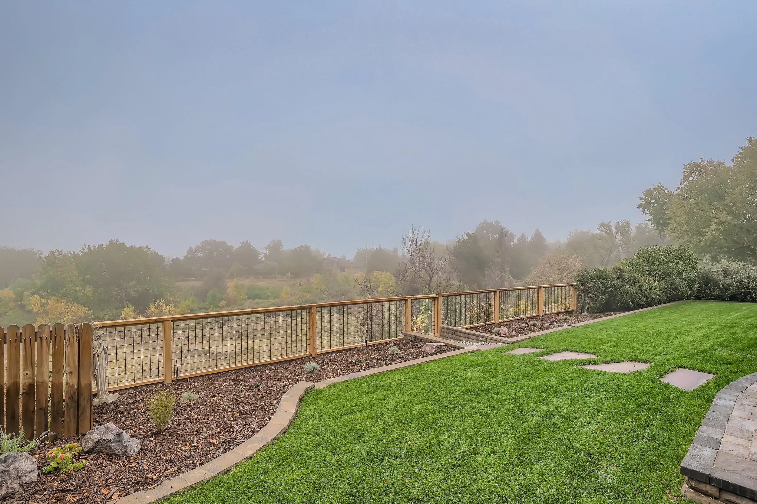 A backyard with a well-manicured lawn, stepping stones, a wooden fence, and trees in the background under a hazy sky.