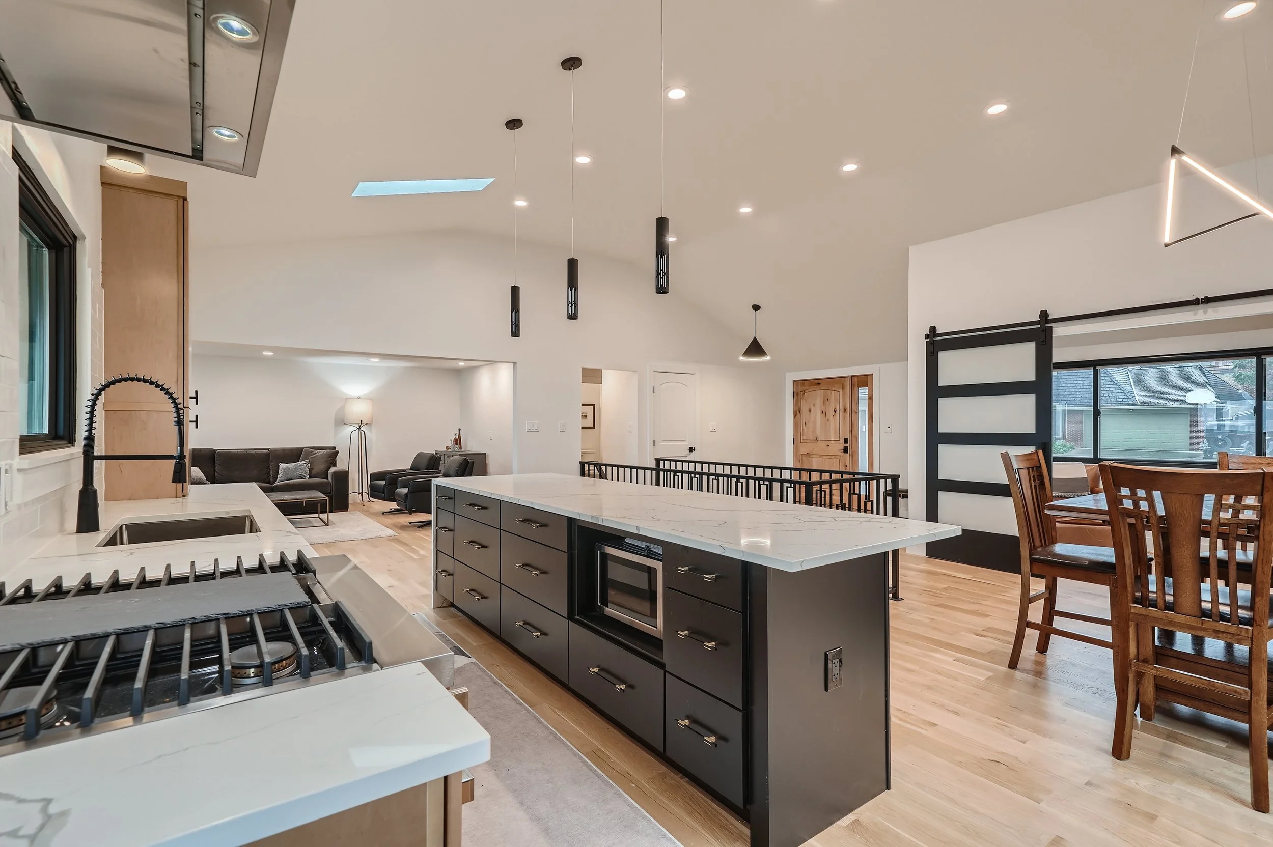 Open-concept kitchen with black cabinets, white marble countertops, and a kitchen island with a microwave, overlooking a living area with black sofas and a wooden dining table near a sliding barn door.