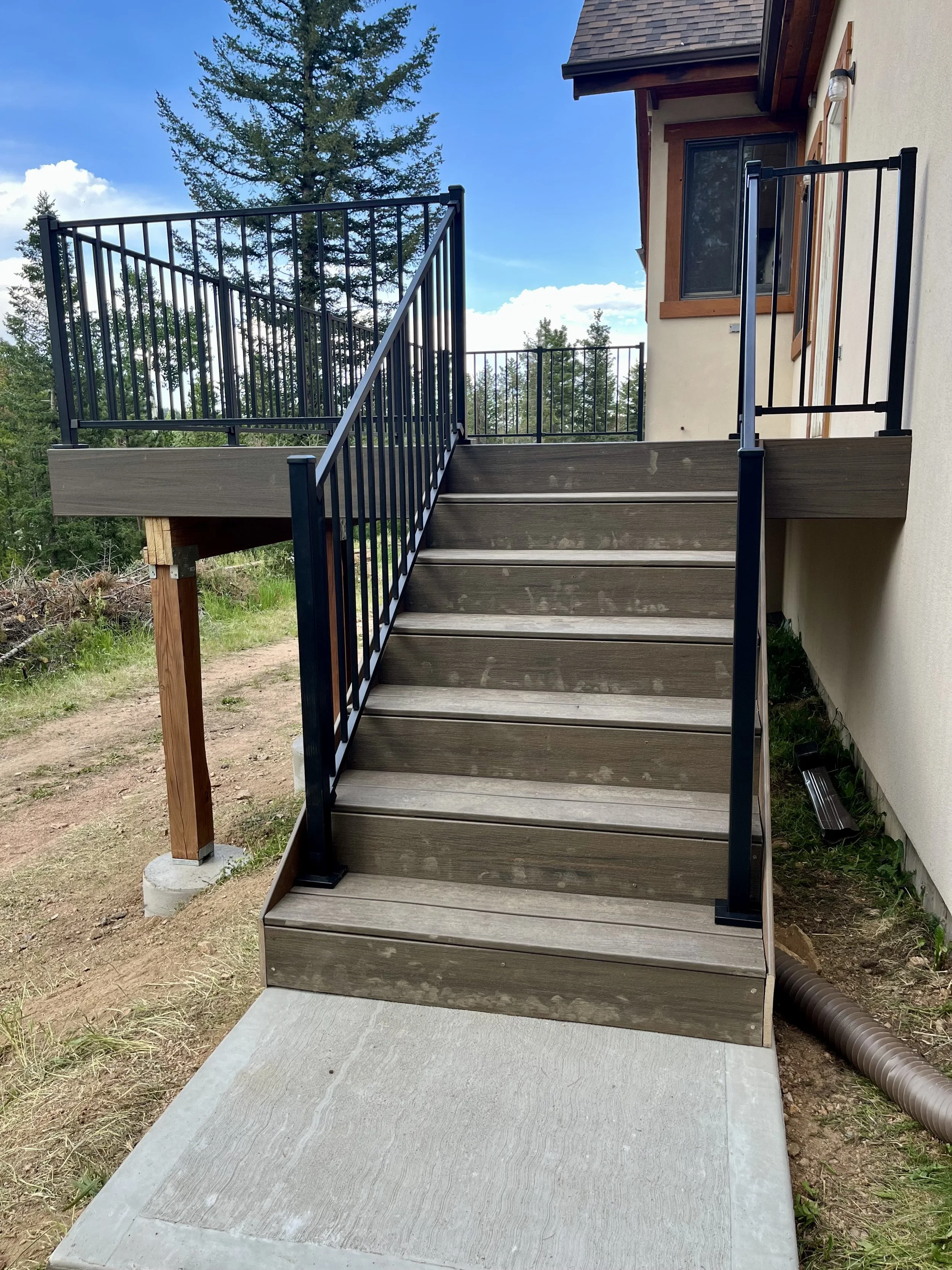 Outdoor staircase with black metal railings leading up to the house, located on a concrete slab, with trees and blue sky in the background.