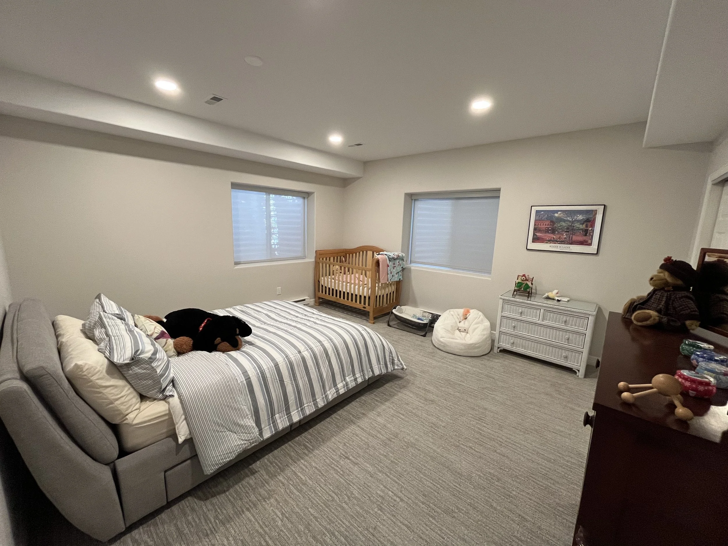A bedroom with a bed, baby crib, and various stuffed animals and toys, illuminated by ceiling lights and window light.