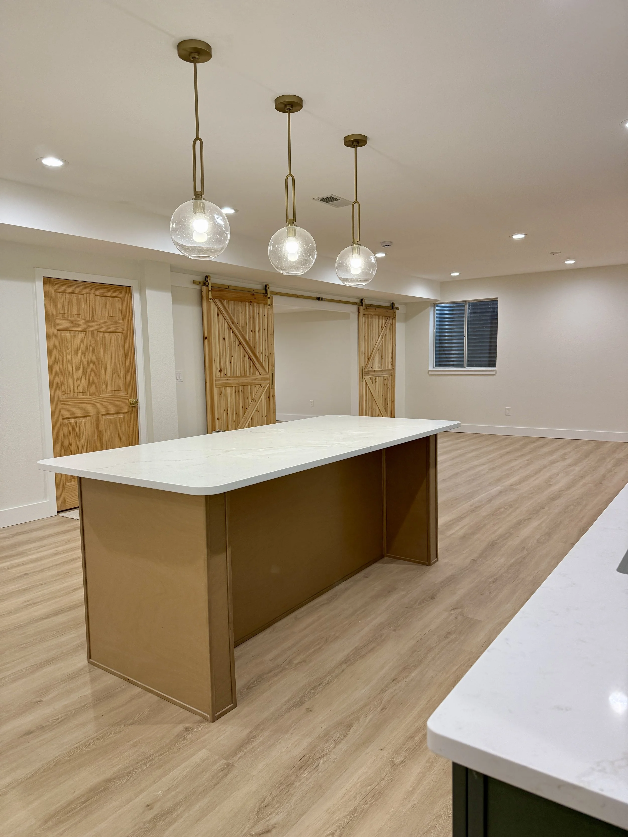 Empty kitchen with a large island with a white countertop, three hanging pendant lights, wooden sliding barn doors, a wooden door, a window with blinds, and light wood flooring.