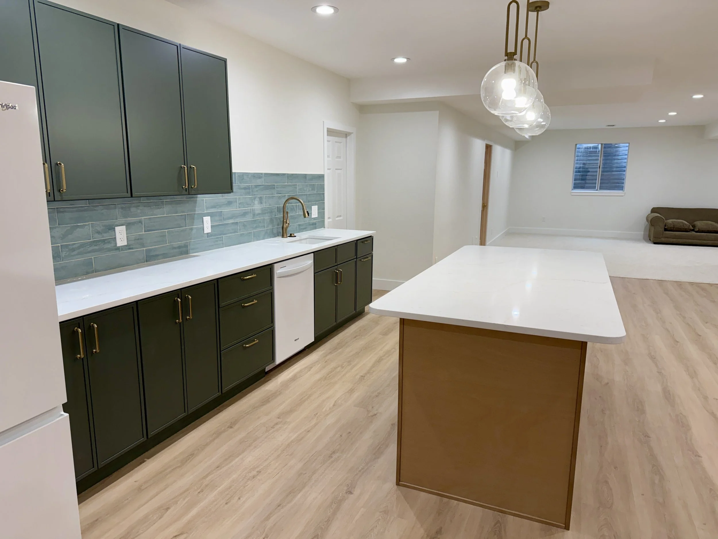 Modern kitchen with dark green cabinets, white countertops, a white sink with a gold faucet, a light wood kitchen island, pendant lights, and a view of the living room with a brown couch and window.