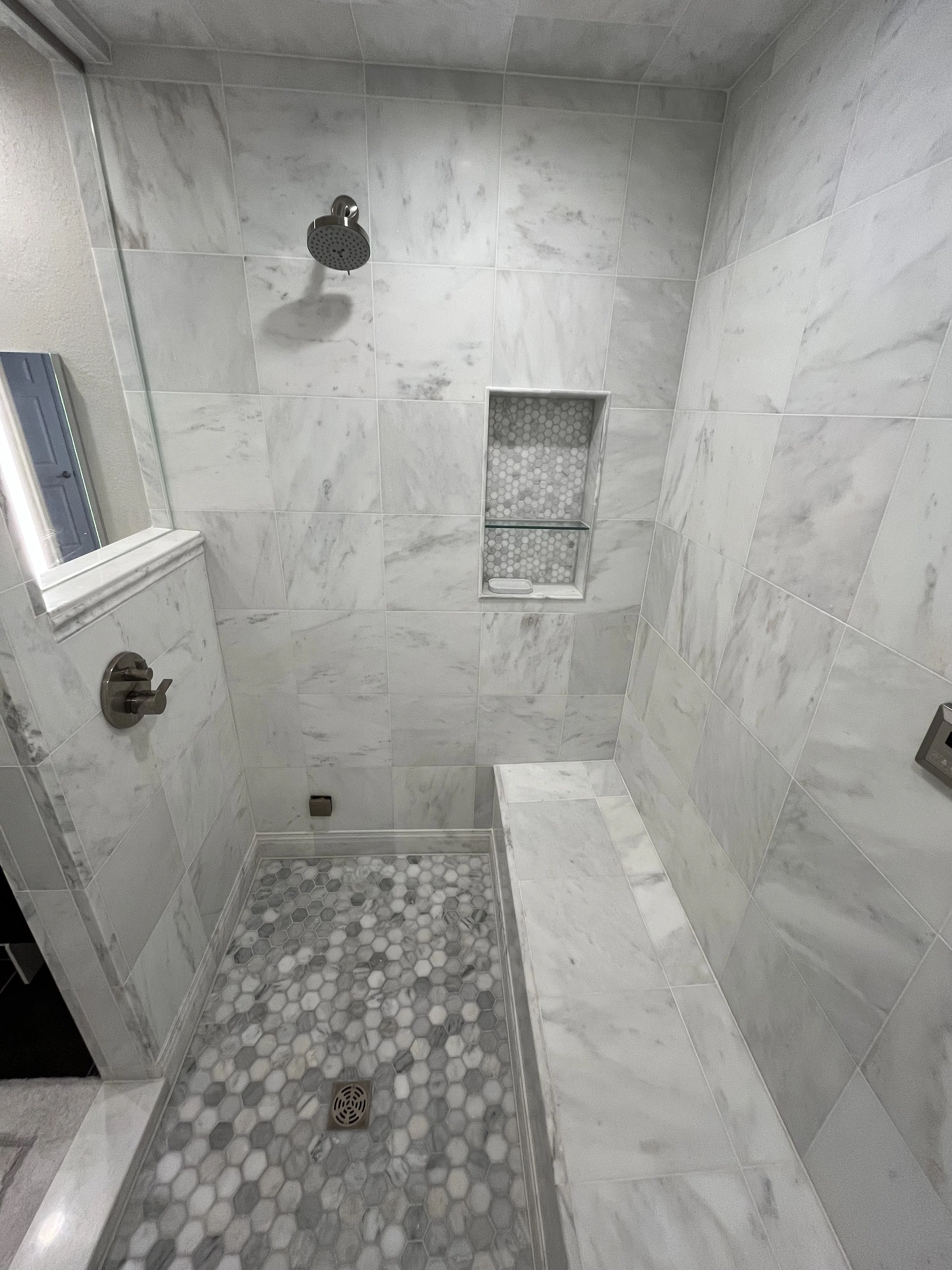 A modern walk-in shower with white marble tiles, a rainfall showerhead, a built-in niche with a glass shelf, a soap dish, and a mosaic tile floor with a drain.